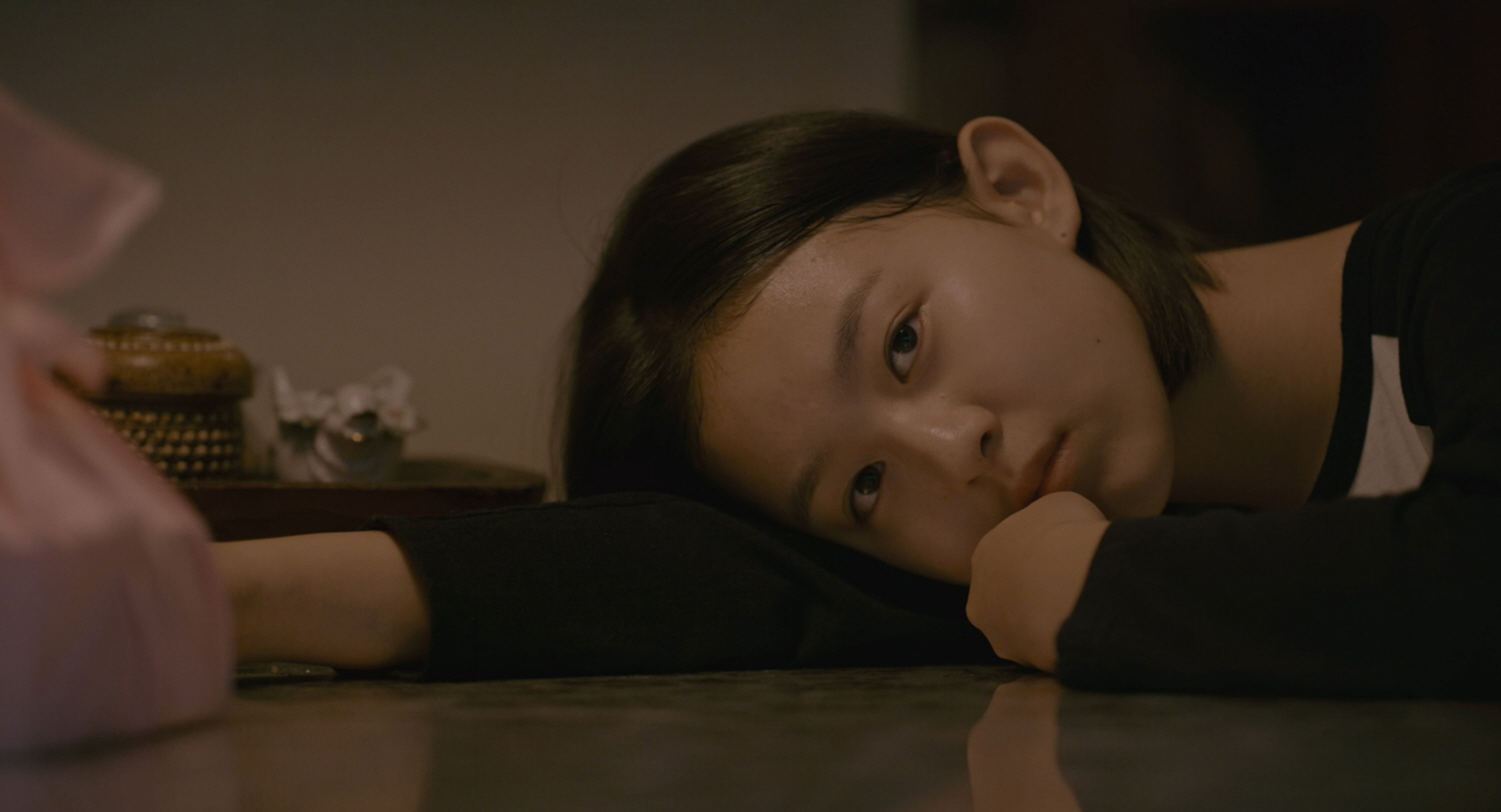A young woman lying on the floor with her head resting on her arm, gazing toward someone nearby.