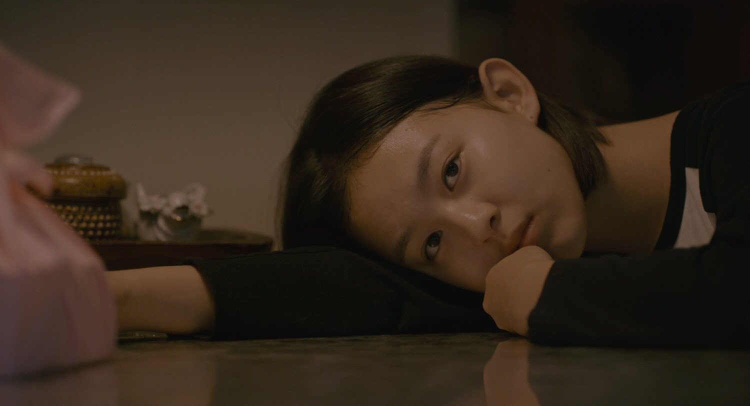A young woman lying on the floor with her head resting on her arm, gazing toward someone nearby.
