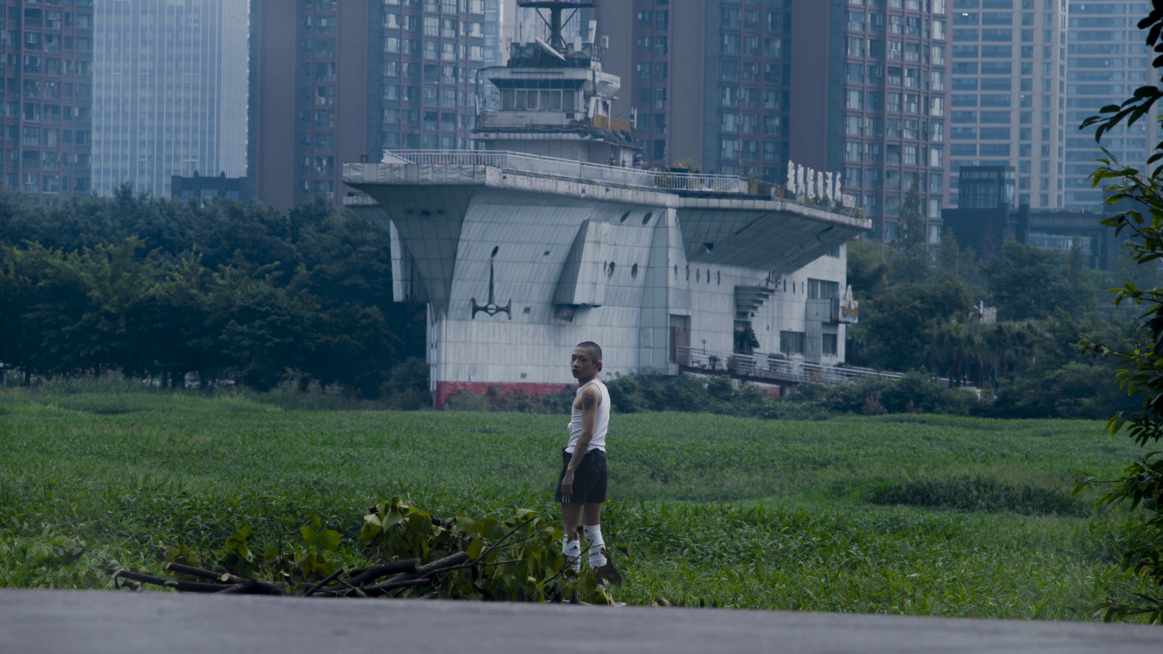 A person standing alone in a green field, with a large concrete structure and high-rise buildings in the background.