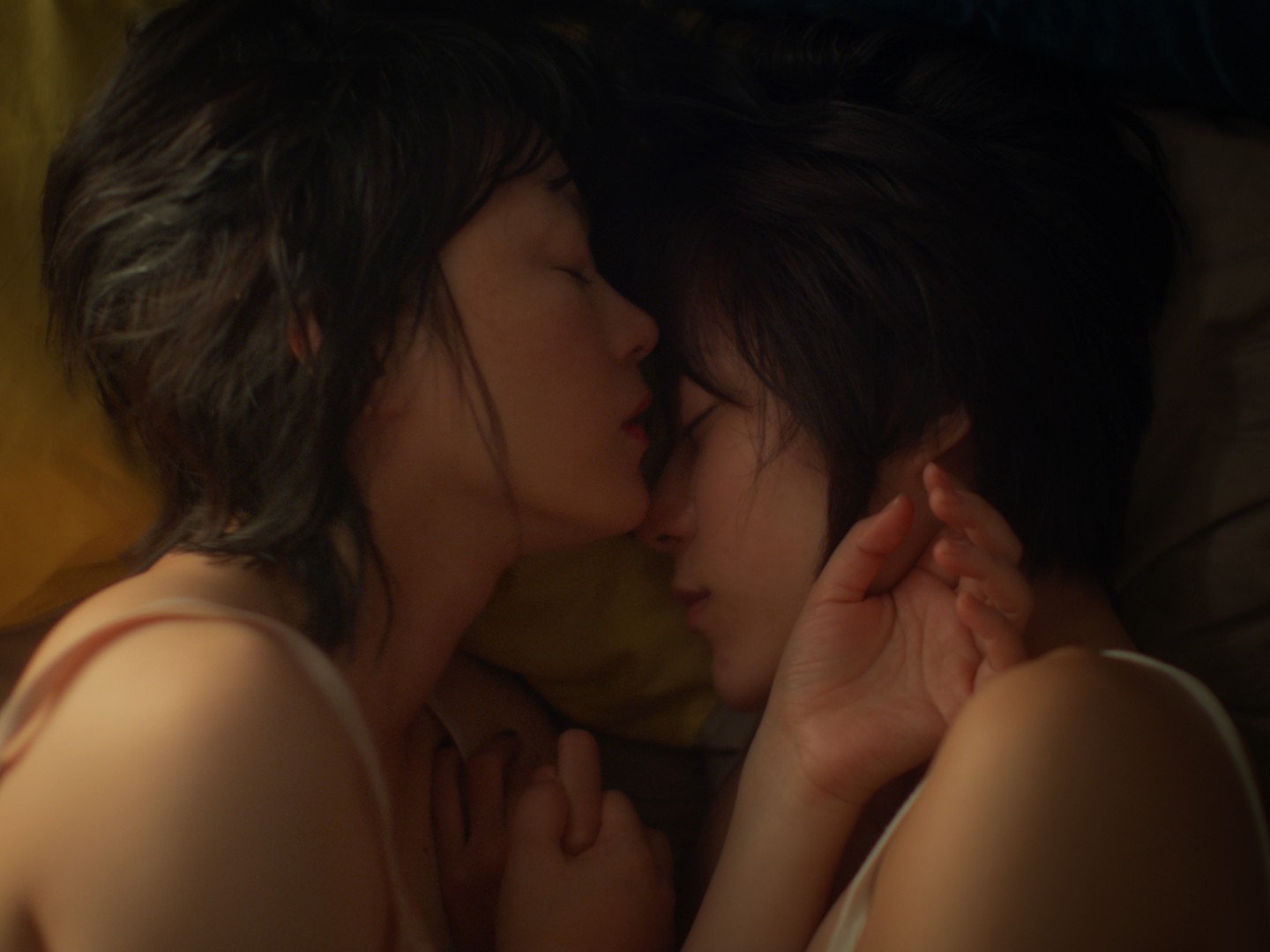 Two women lying closely together in bed, one gently touching the other’s face.