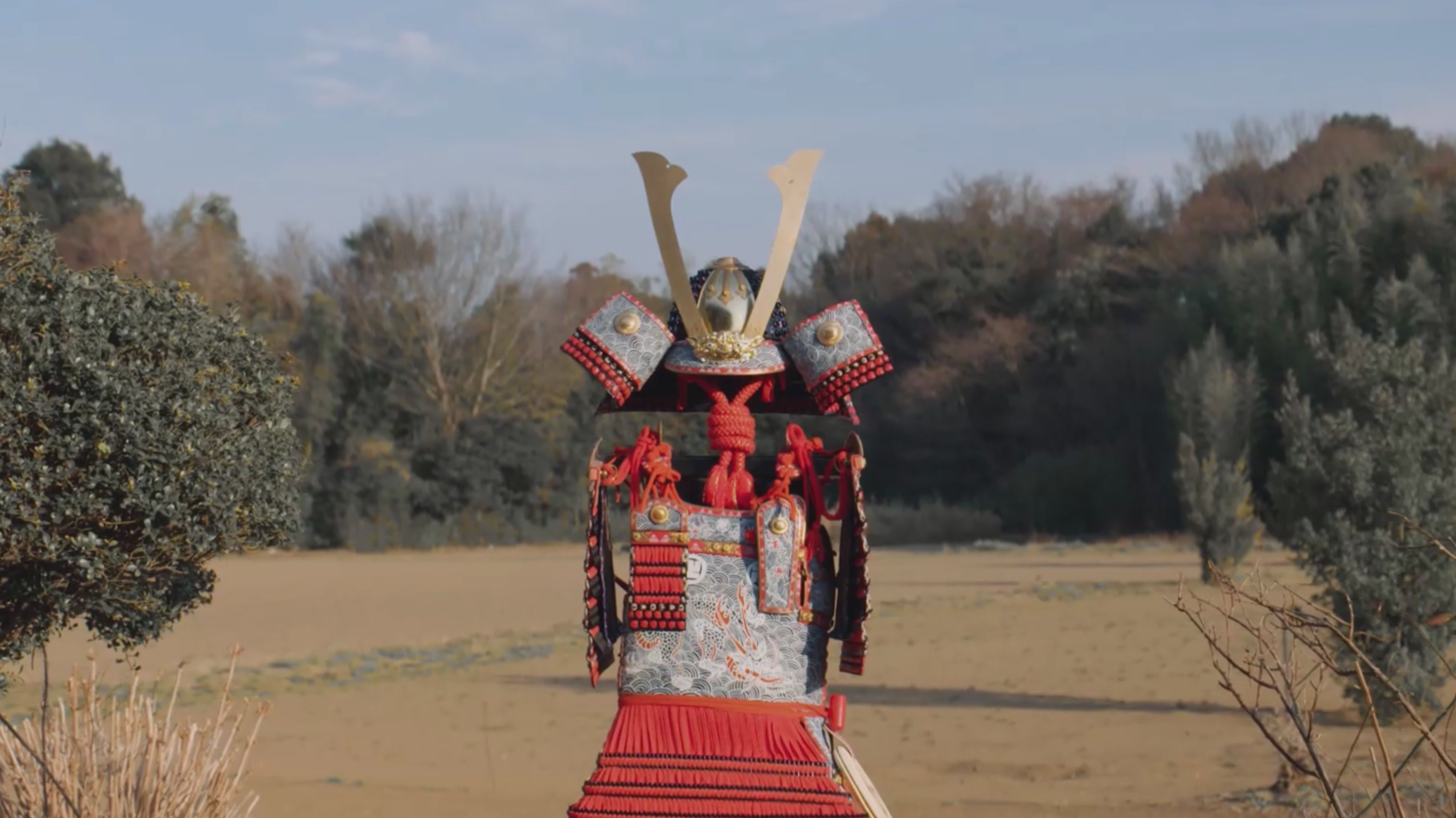 A figure in ornate red and silver samurai armour standing alone in an open landscape.