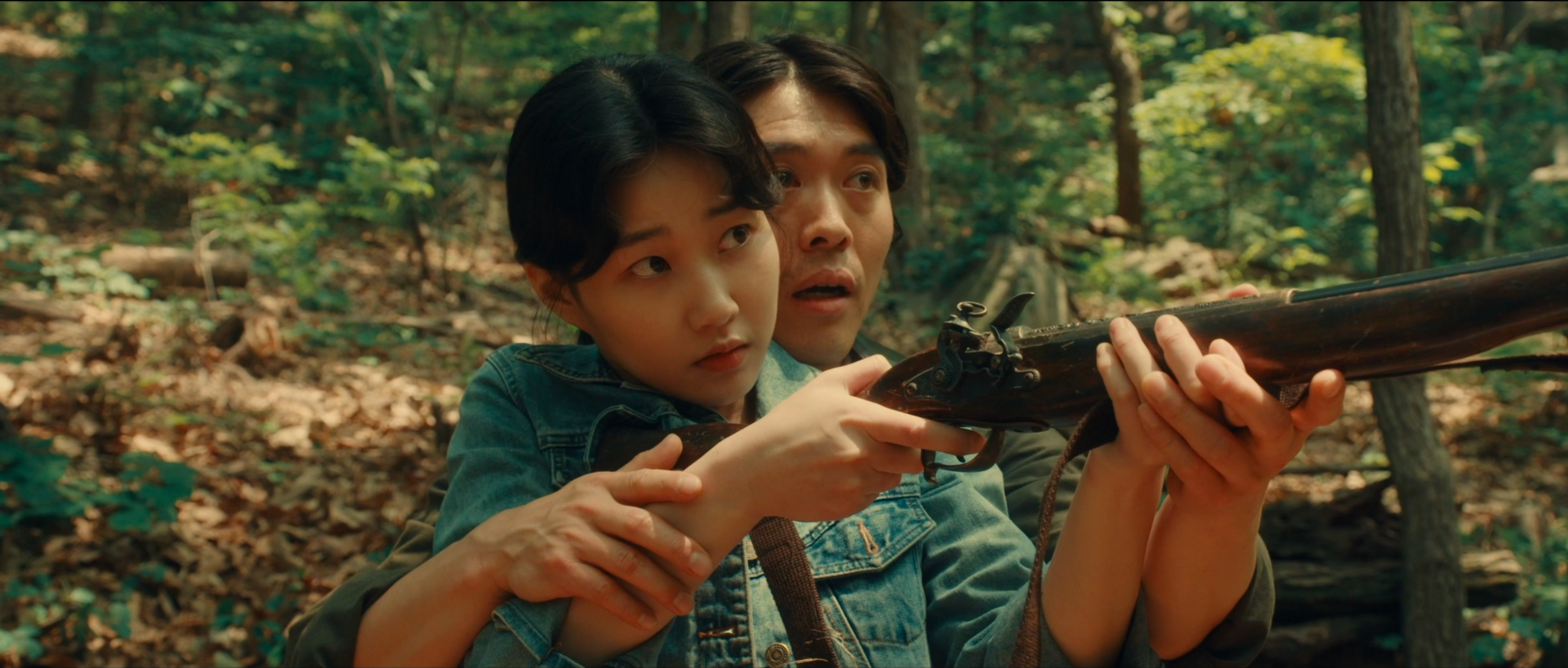 Two people in a forest, with one standing behind the other and guiding their hands as they hold a rifle.