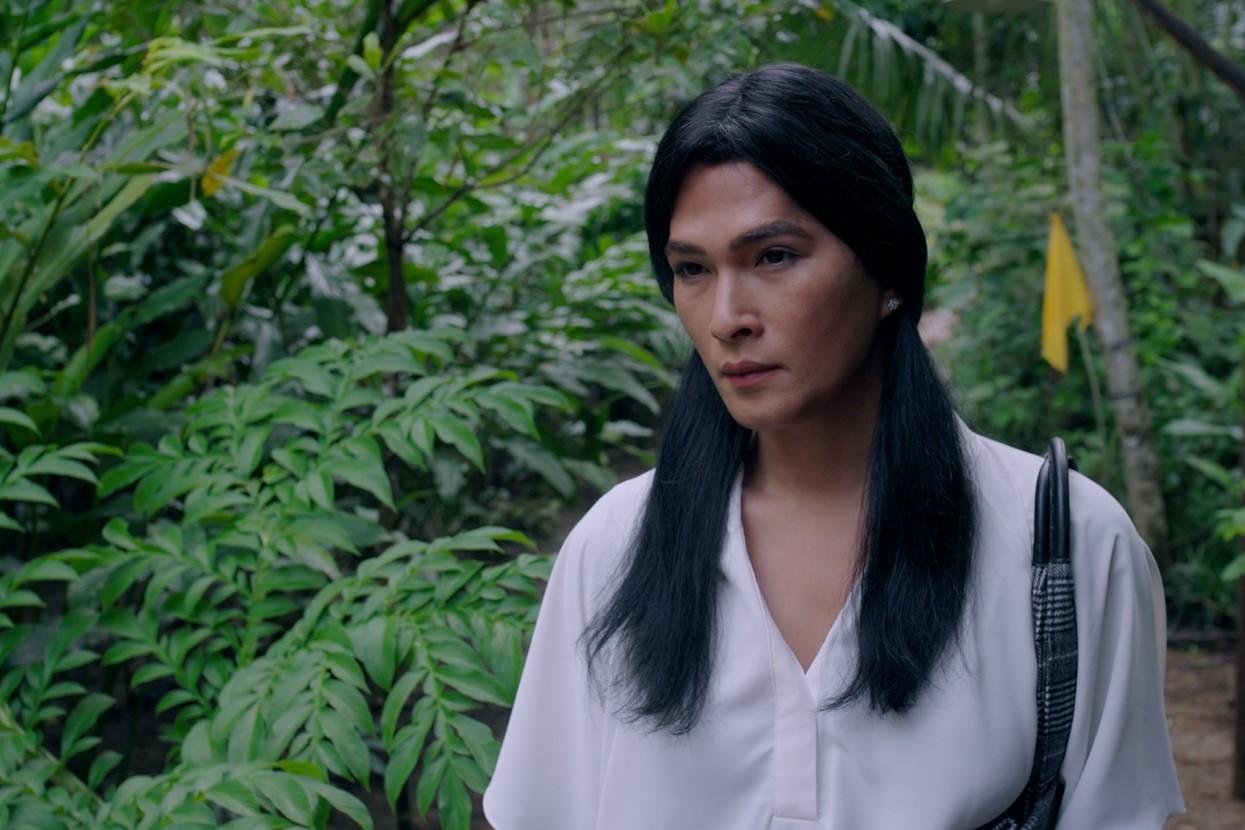 A woman with long dark hair standing on a path surrounded by dense green foliage.