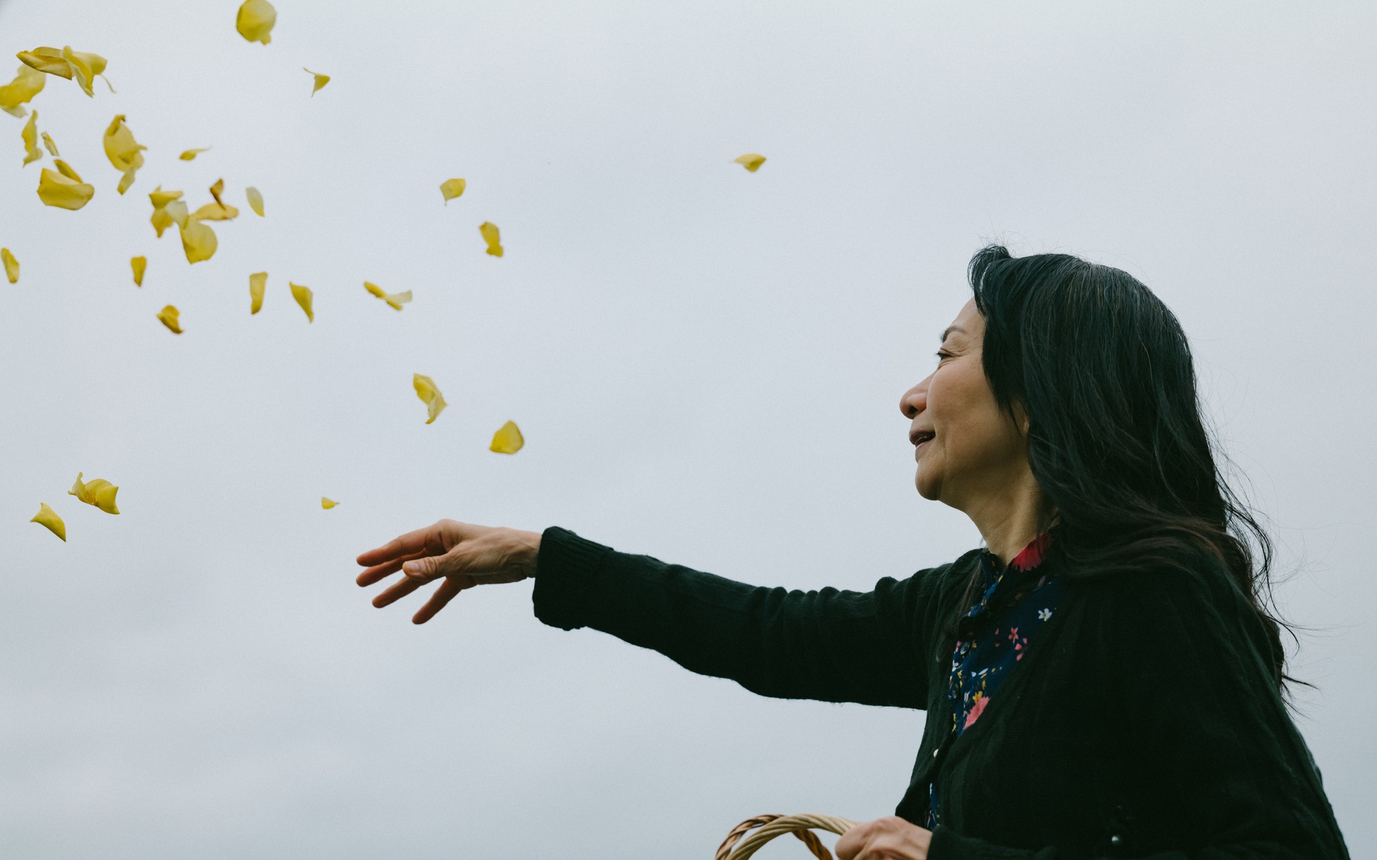 A woman scattering yellow flower petals into the air against an overcast sky.