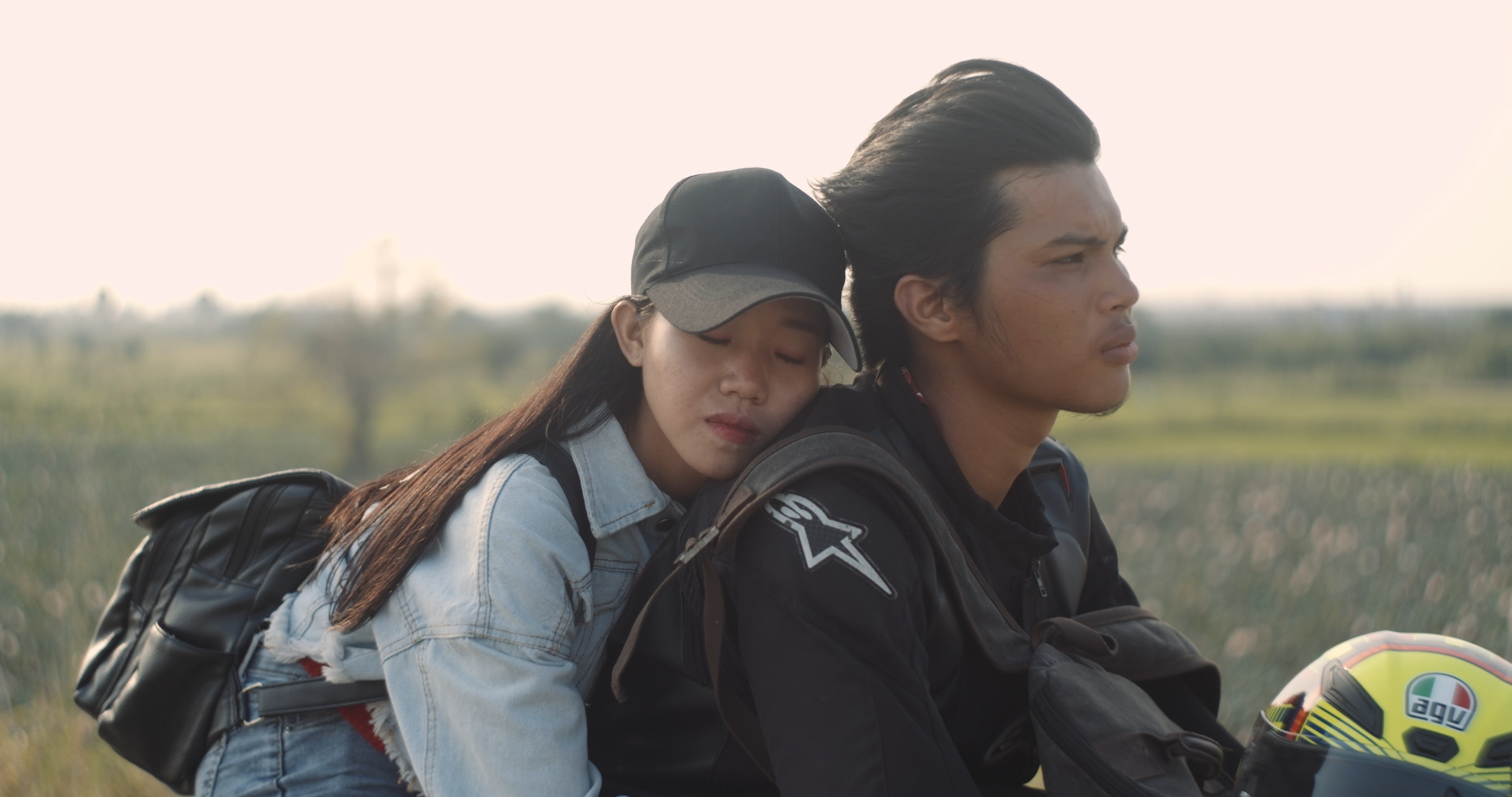A woman resting with her eyes closed against a man seated on a motorcycle in an open rural landscape.