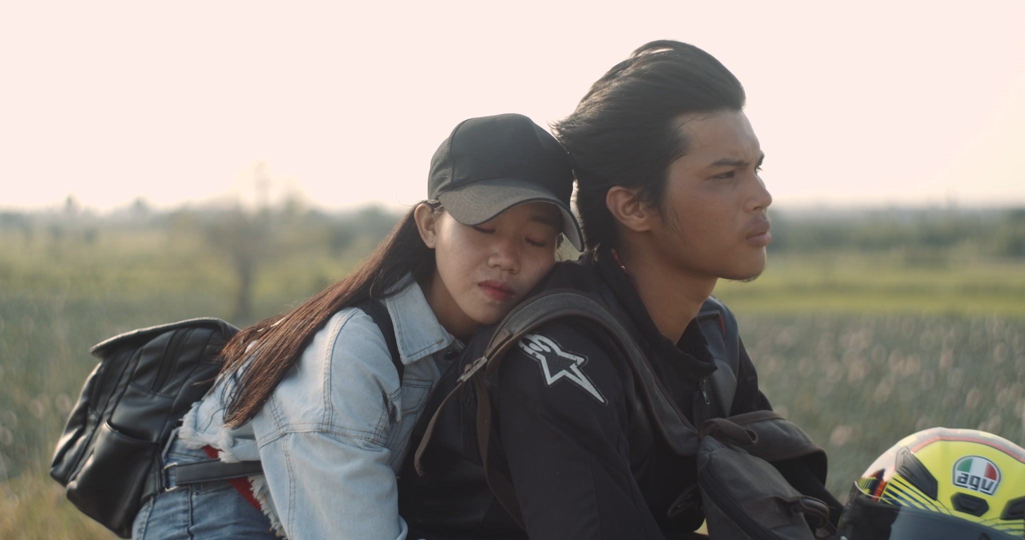 A woman resting with her eyes closed against a man seated on a motorcycle in an open rural landscape.