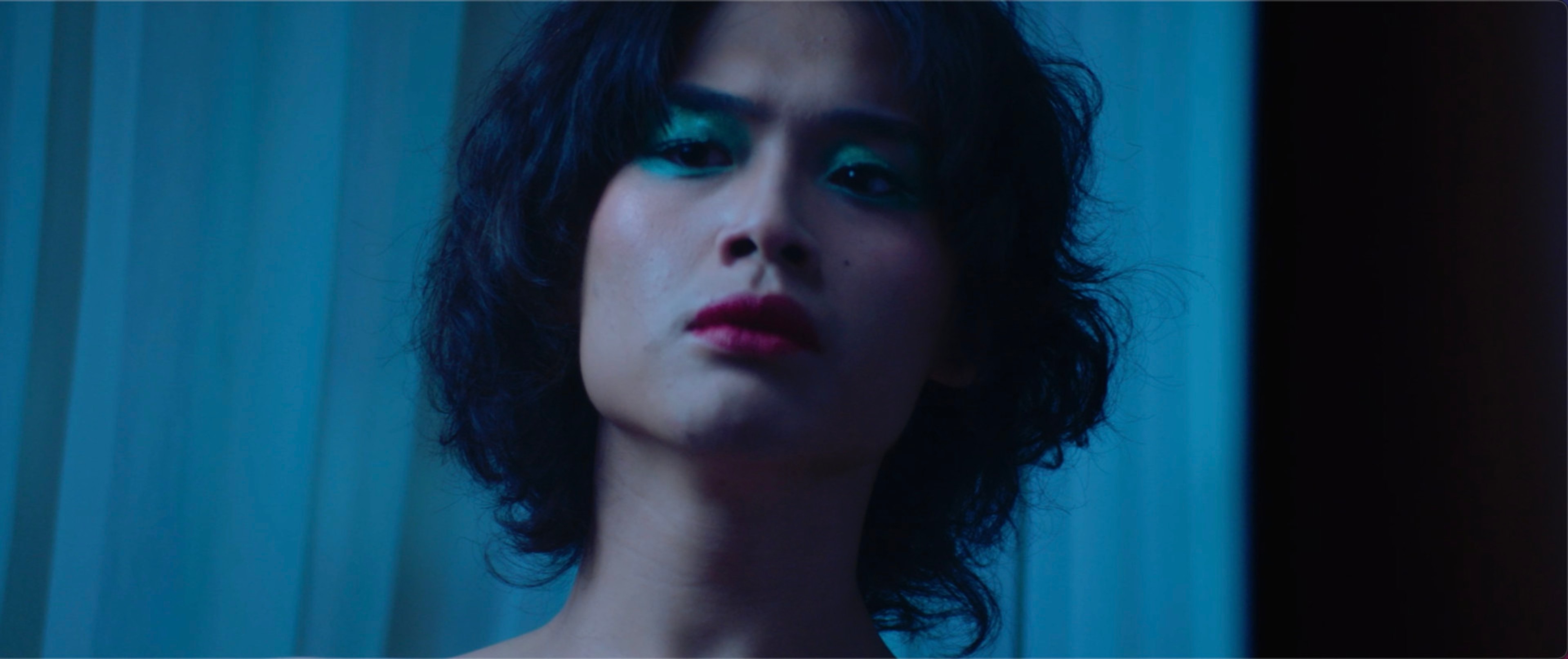 A close-up of a person with dark wavy hair, bold eye makeup and red lipstick under cool blue lighting.