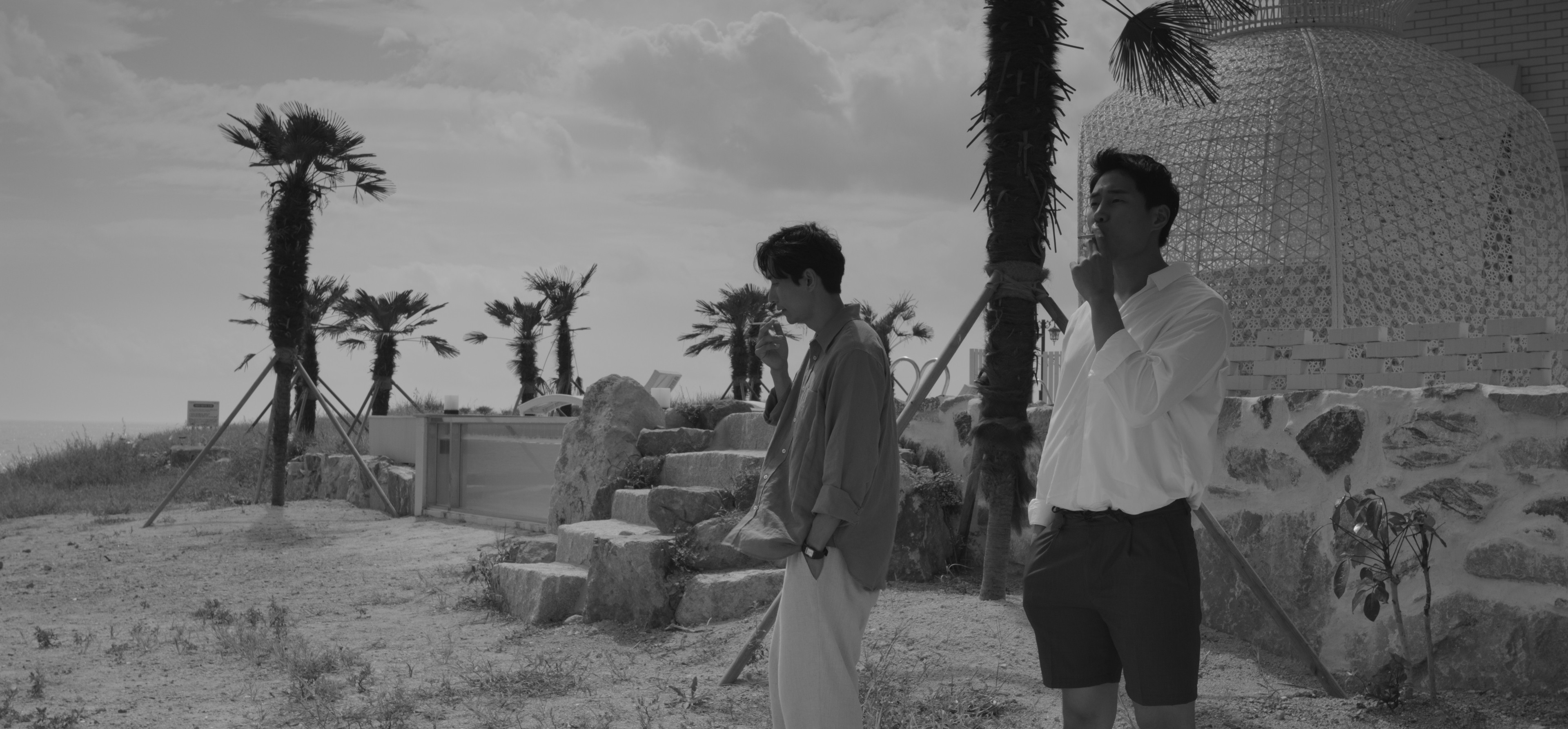 Two men standing apart in a coastal setting with palm trees, shown in black and white.