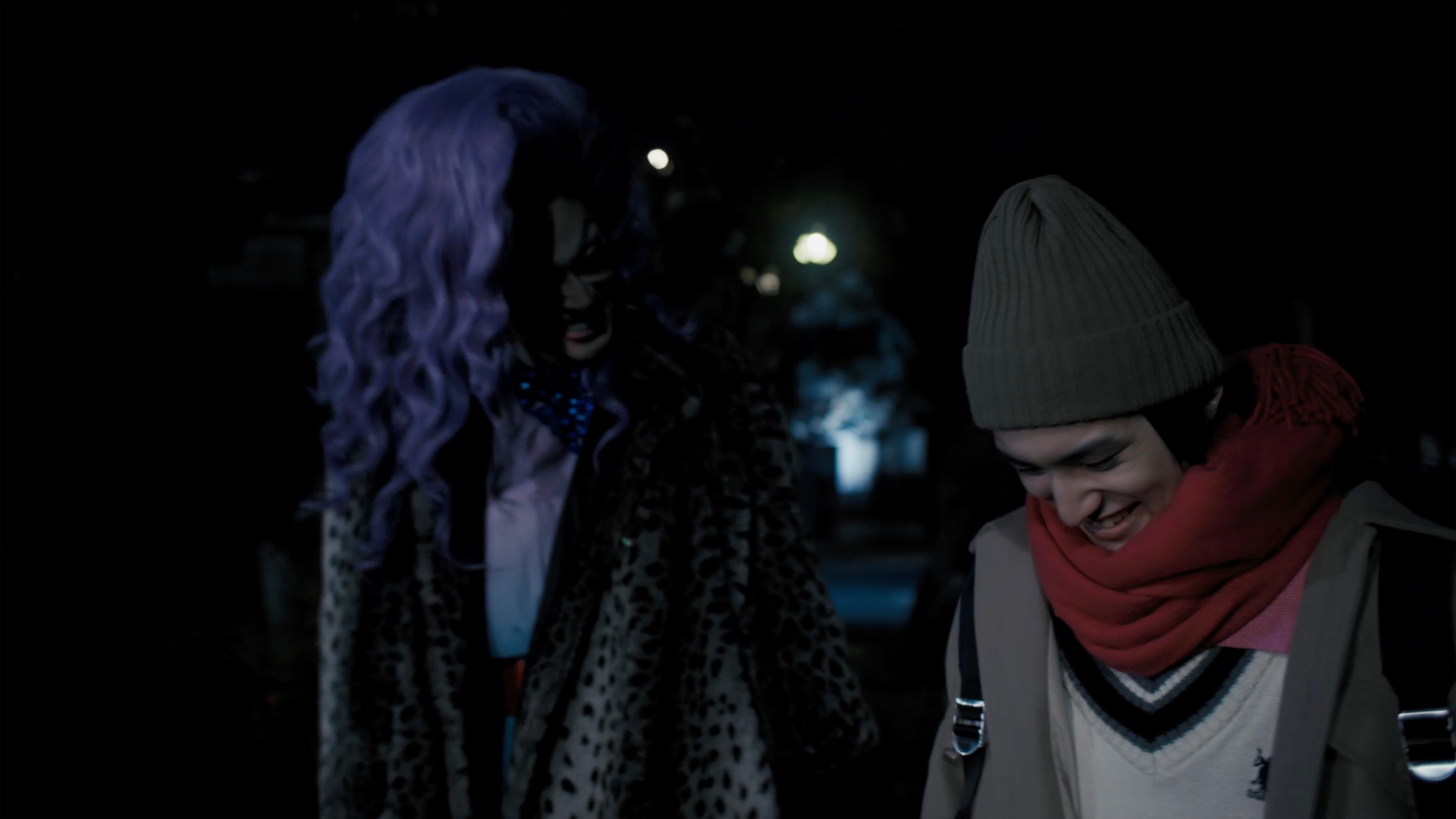 Two people walking together at night, one wearing a colourful wig and patterned coat while the other smiles in a beanie and scarf.