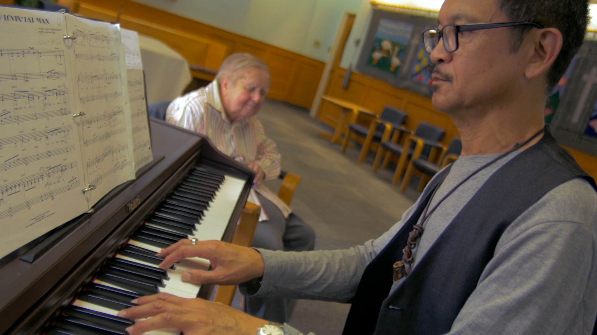 A man playing piano from sheet music while another person sits nearby listening.