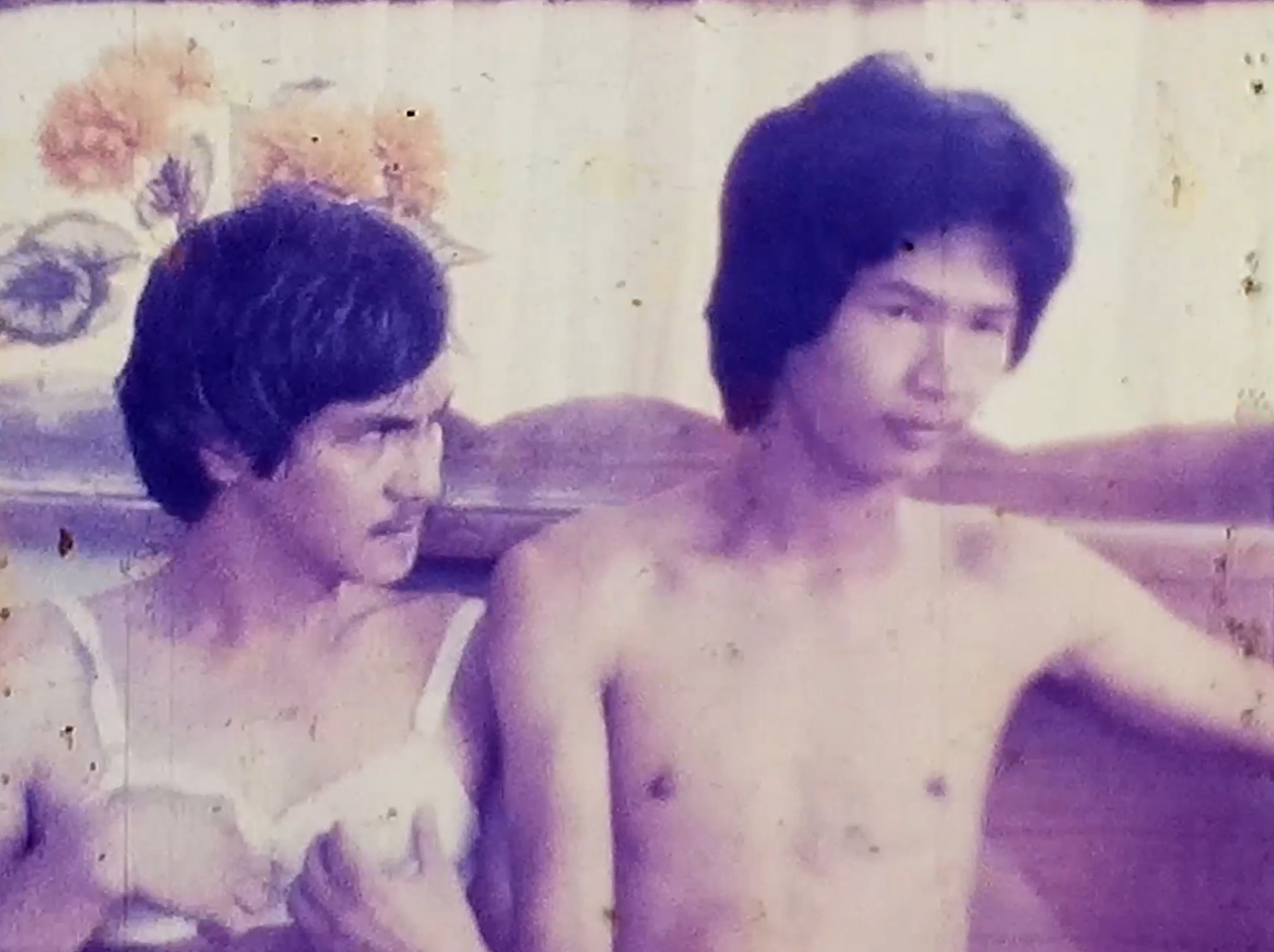 Two young men sitting closely together, one shirtless, in a faded vintage film still.