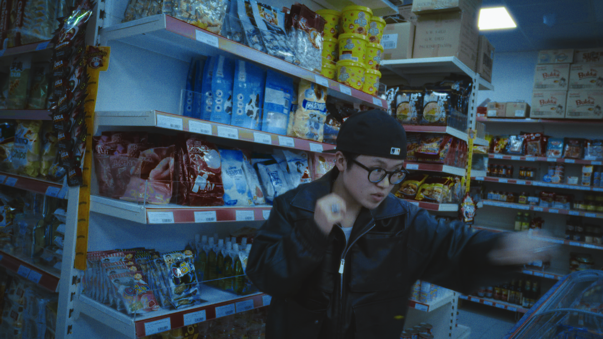 A young person wearing glasses and a backwards cap standing in the aisle of a convenience store.