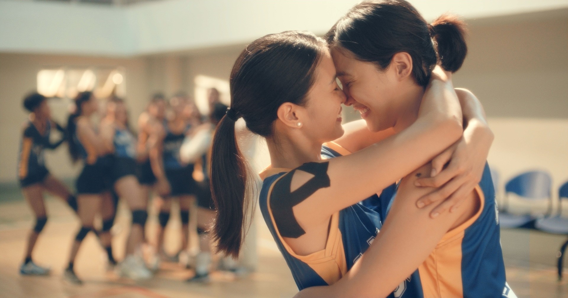 Two women in sports uniforms embracing closely in a gym, with teammates celebrating in the background.
