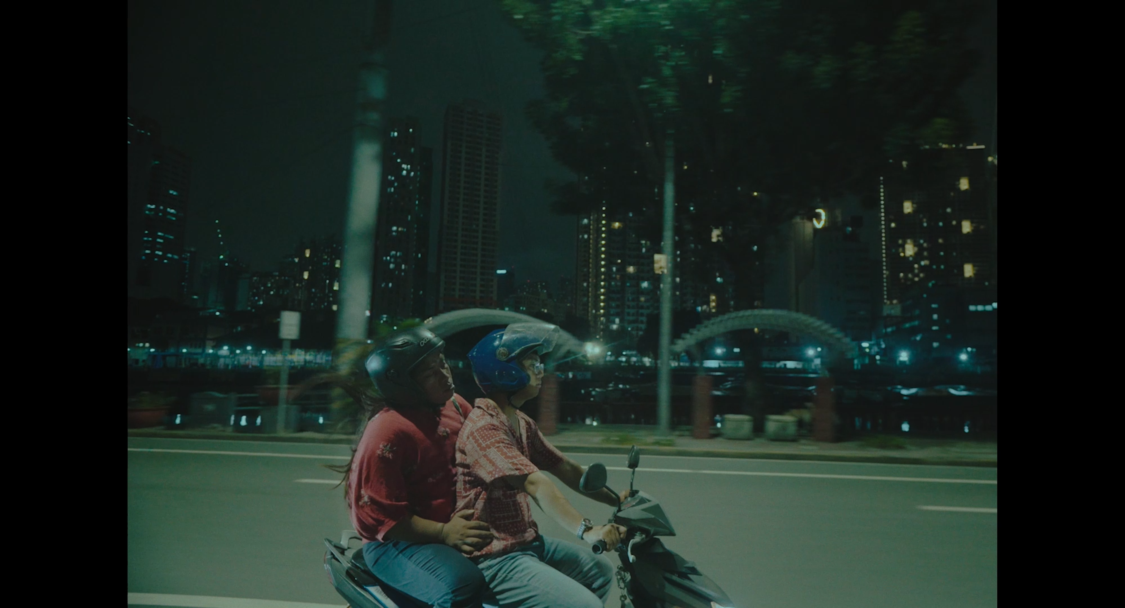 Two people wearing helmets riding together on a scooter through a city at night.