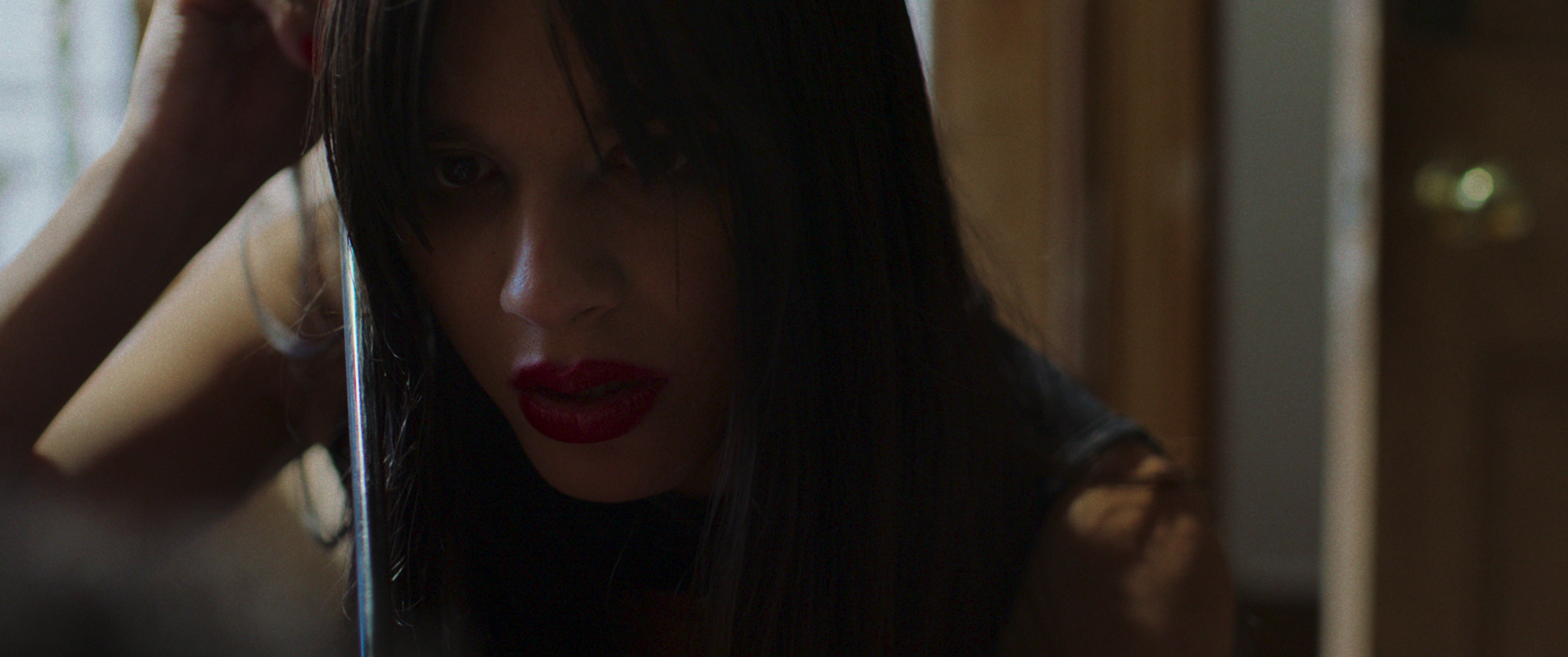 A close-up of a woman with long dark hair and red lipstick looking into a mirror in a dimly lit room.