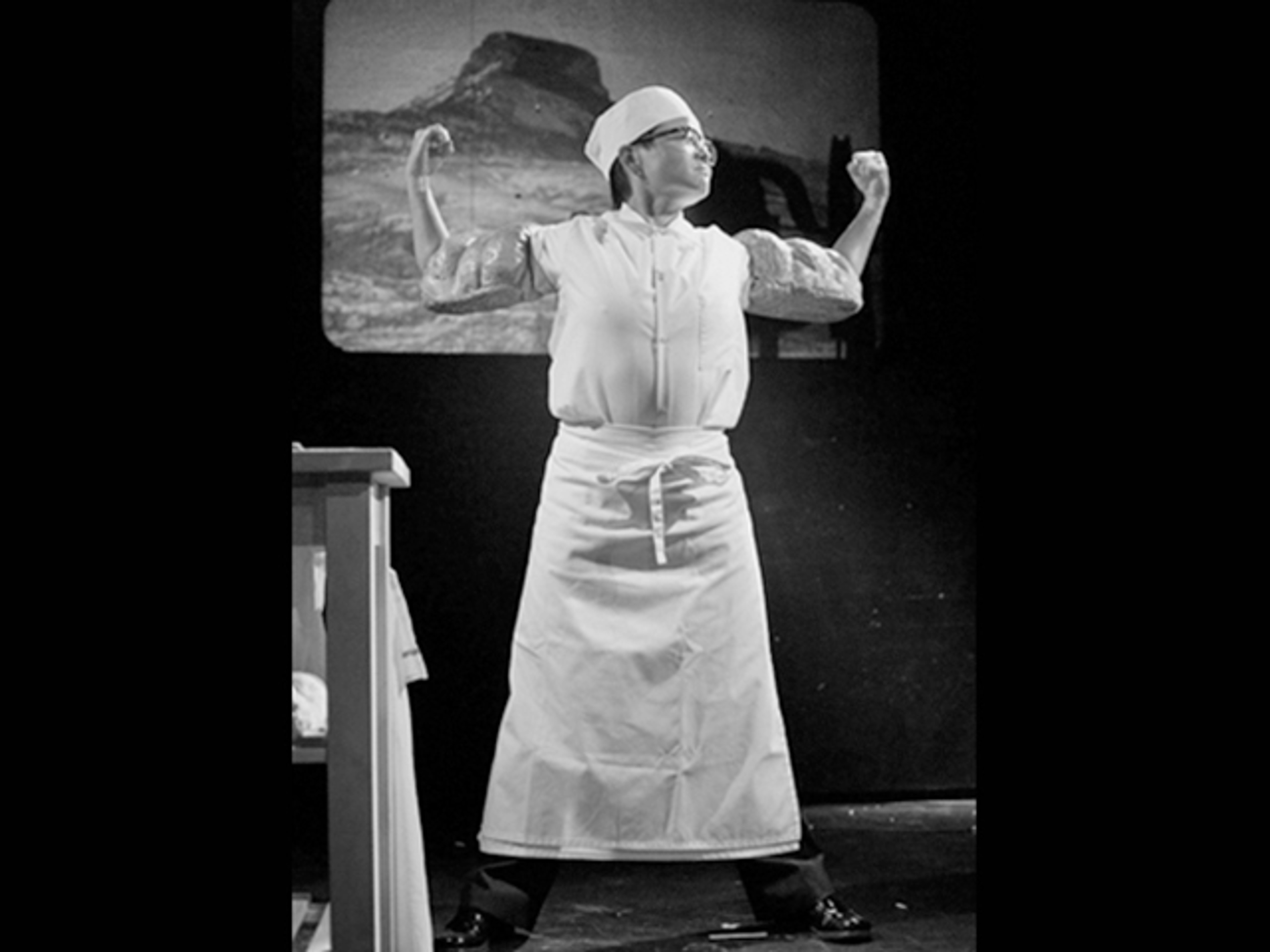 A black-and-white image of a performer in a white costume and head covering, standing with arms raised on a stage.