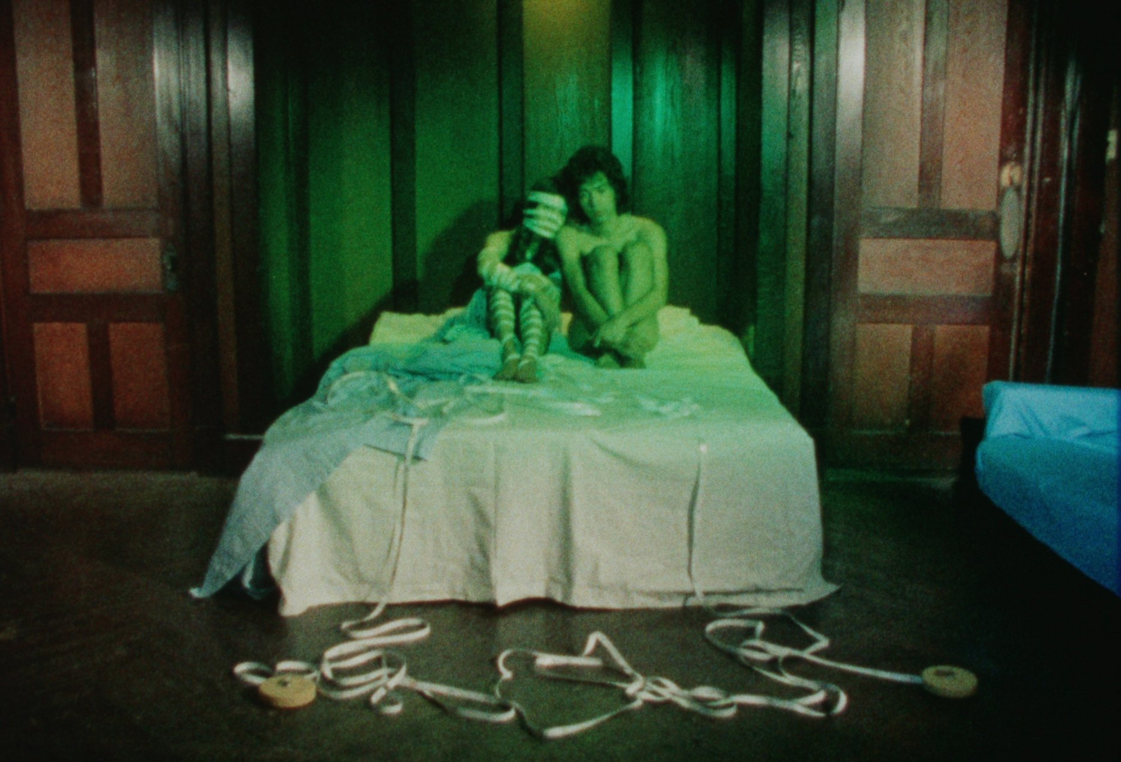 Two people sitting closely together on a bed in a dim, green-lit room.