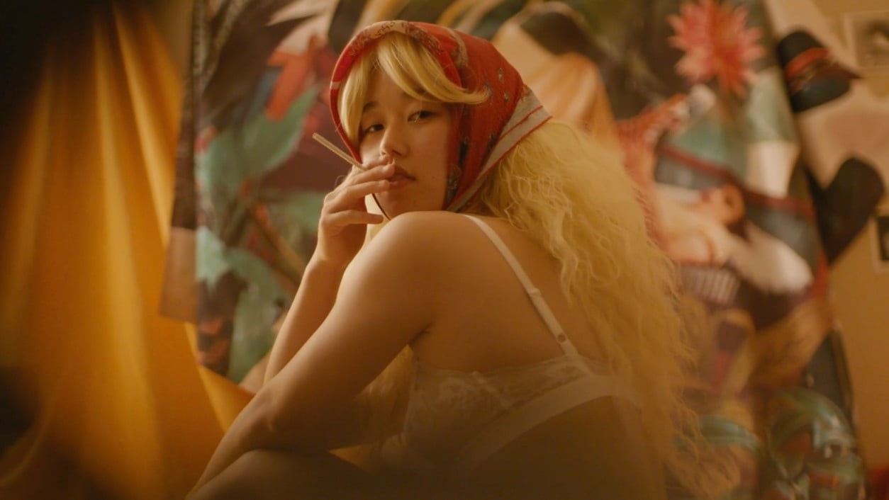 A person with long blonde hair and a red headscarf looking back toward the camera in a warmly lit interior.