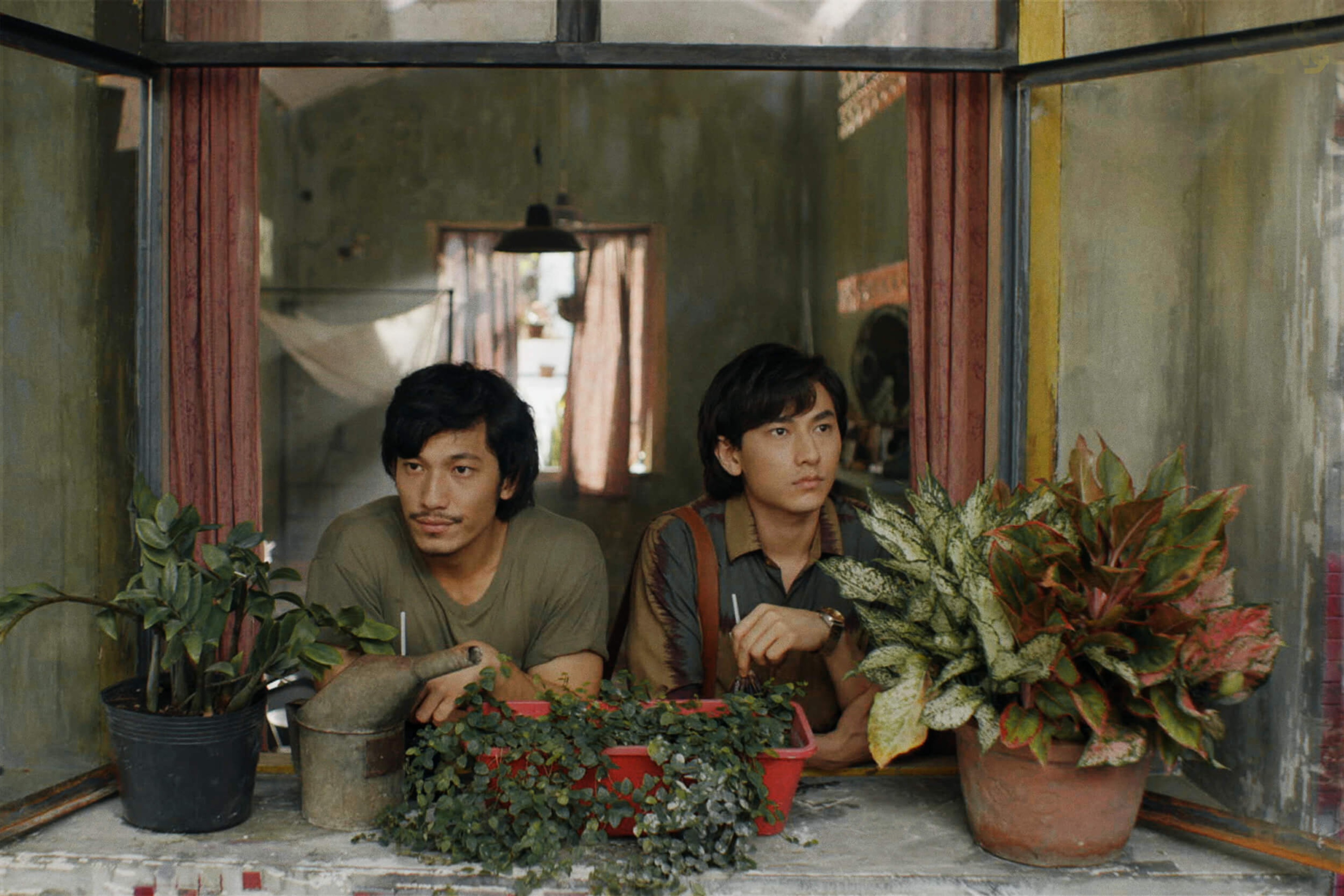 Two men standing next to each other in a window sill with potted plants.