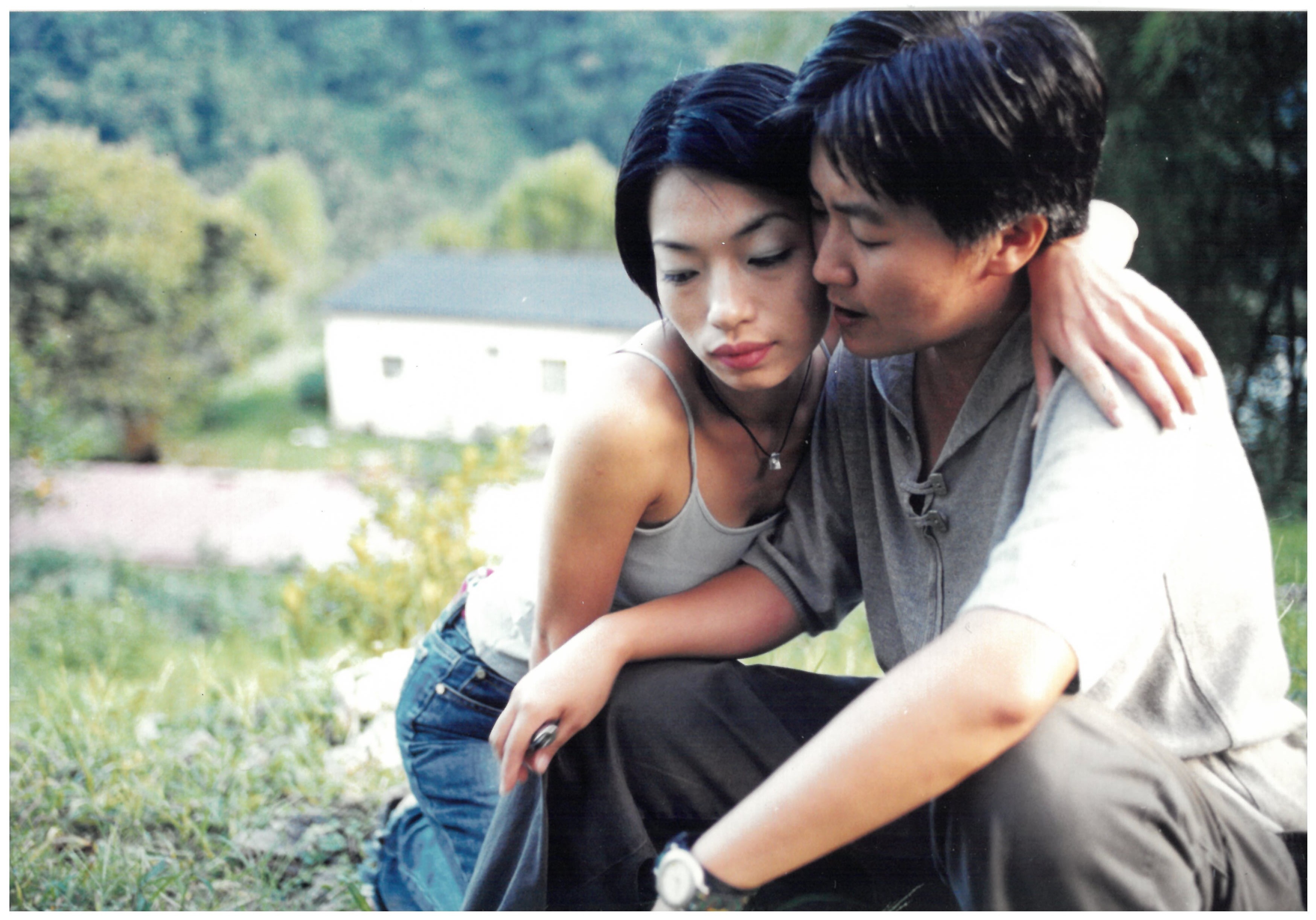 Two people sitting closely together outdoors, one with an arm around the other’s shoulders.