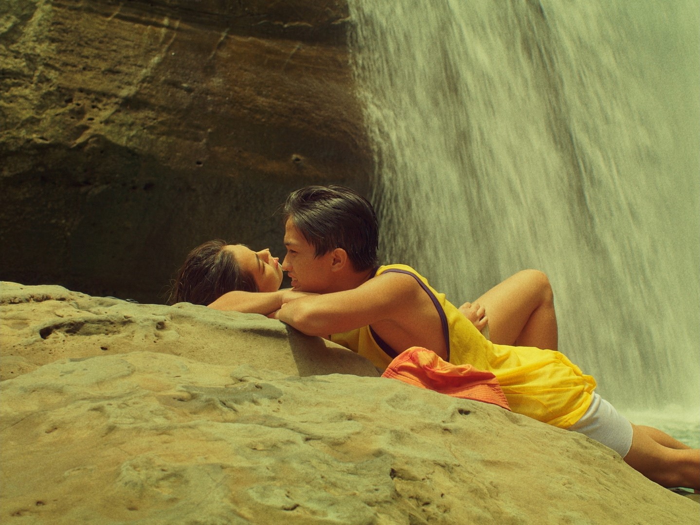 Two people embracing and kissing on rocks beside a waterfall.