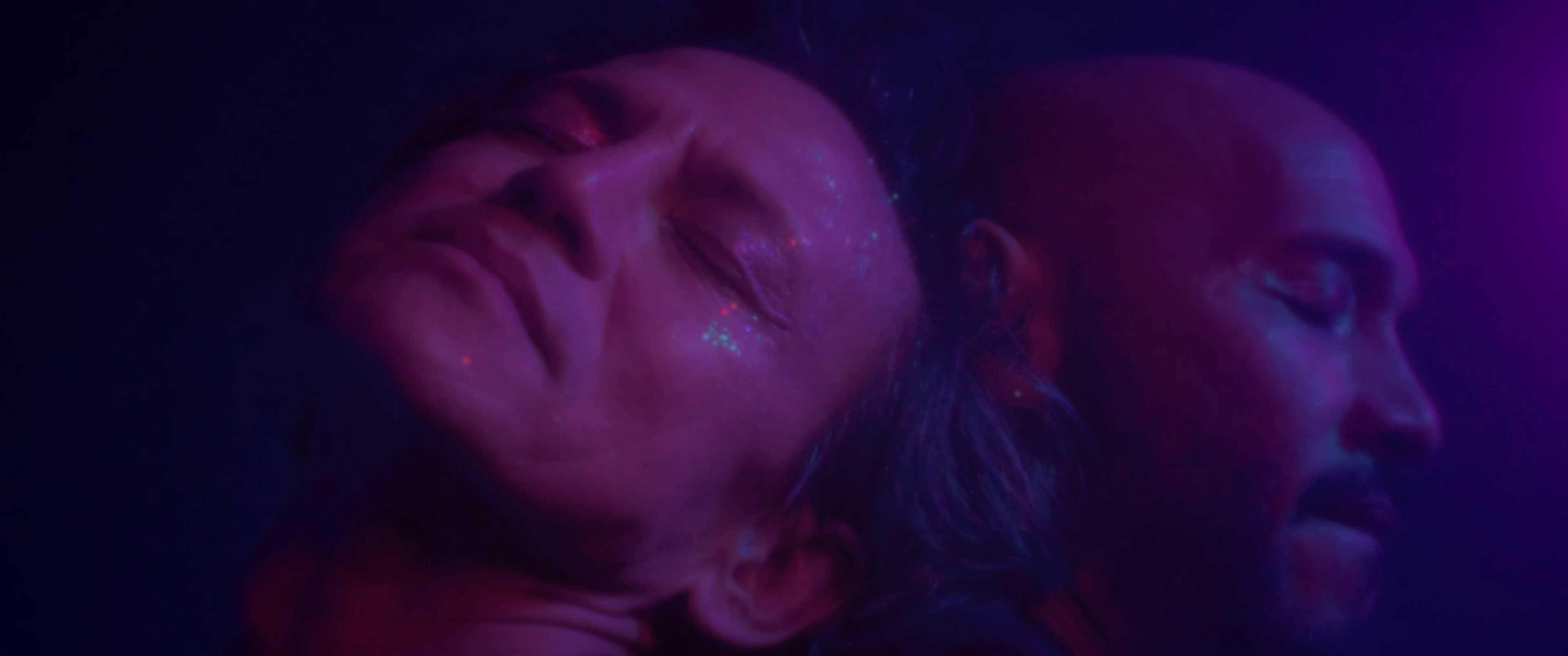 Two faces with closed eyes bathed in pink and purple light.