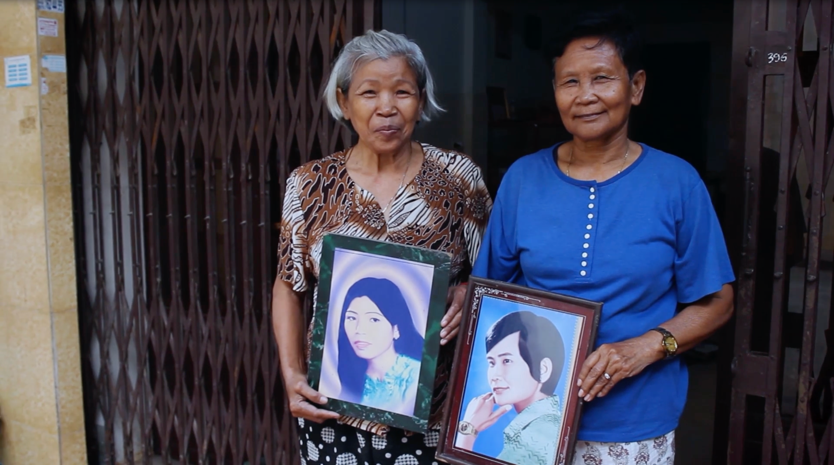 Two figures stand side-by-side each holding a framed portrait.