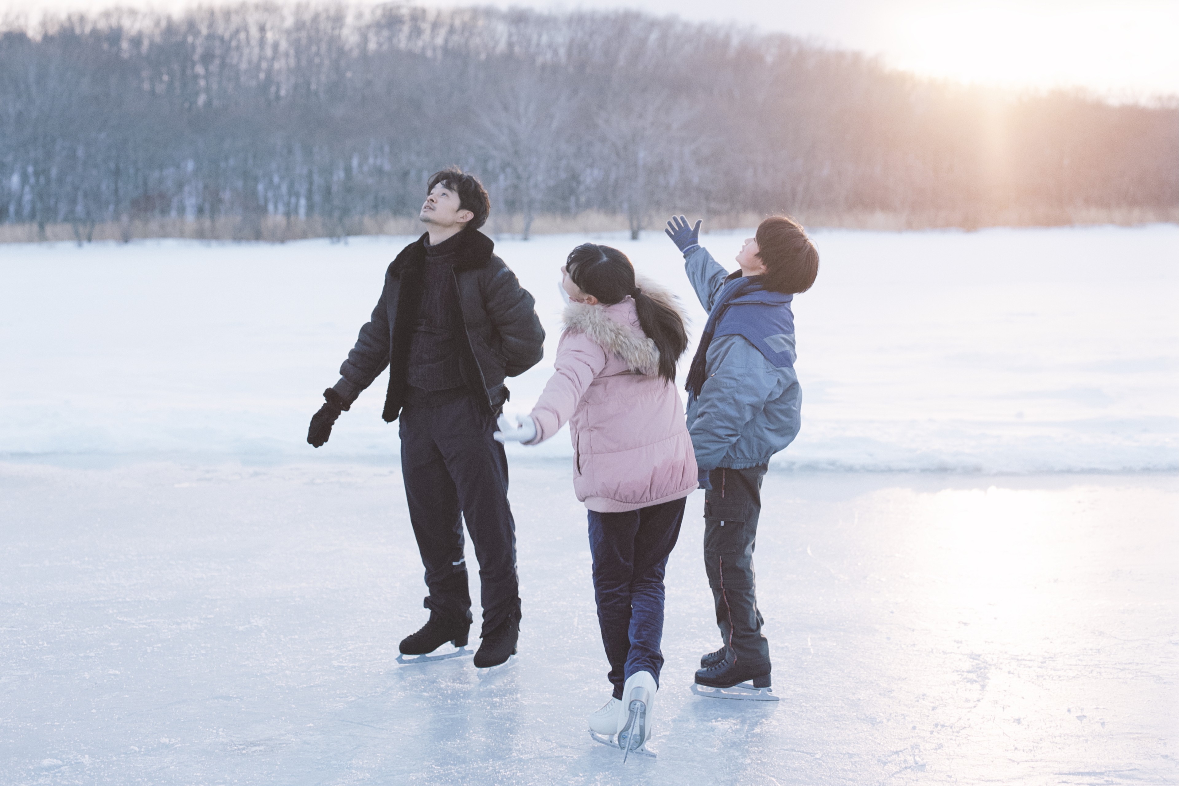 Three people in winter clothing standing together on a frozen lake at sunset.