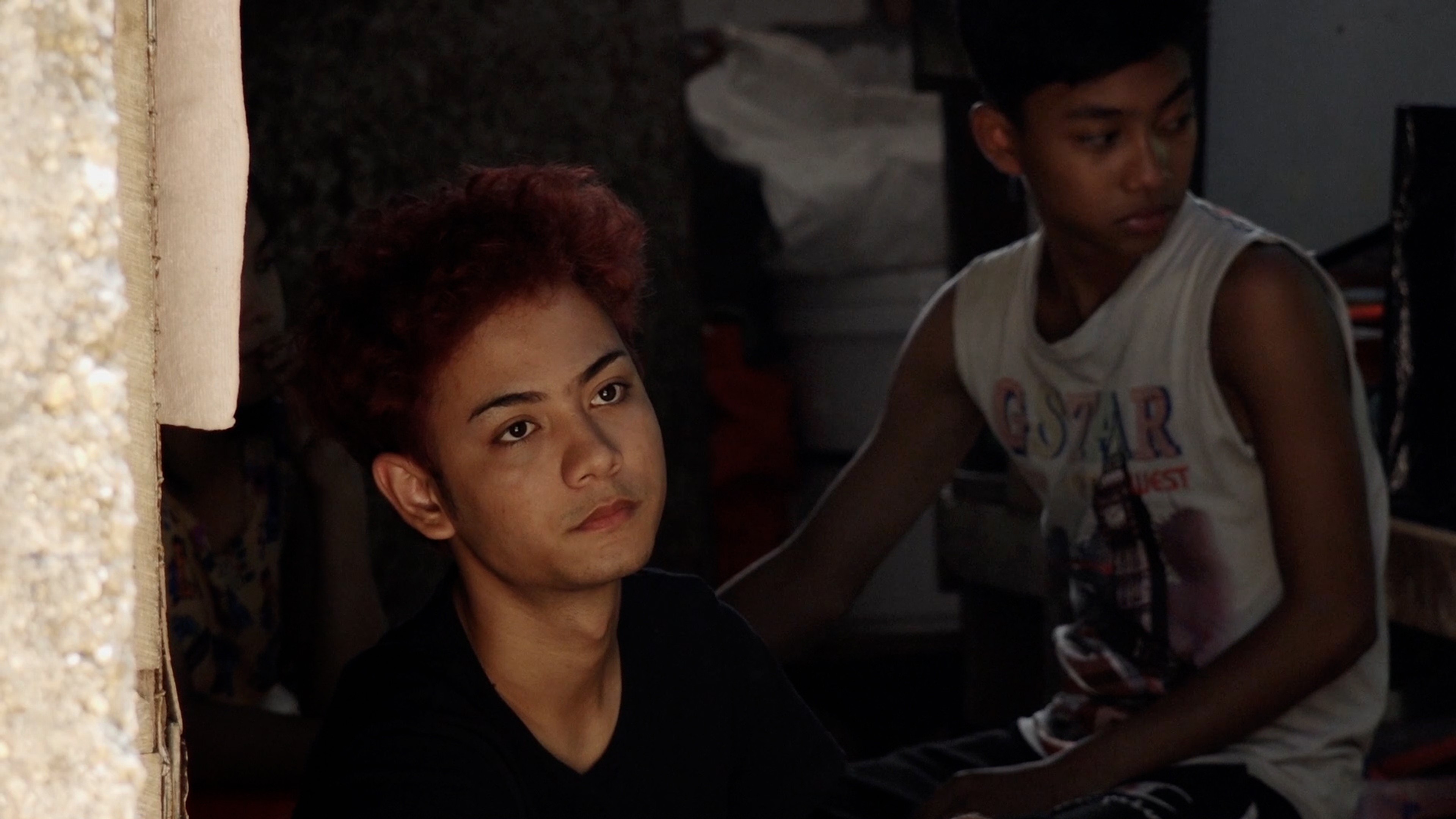 Two young men sitting together in a dim interior, one looking ahead while the other turns to the side.