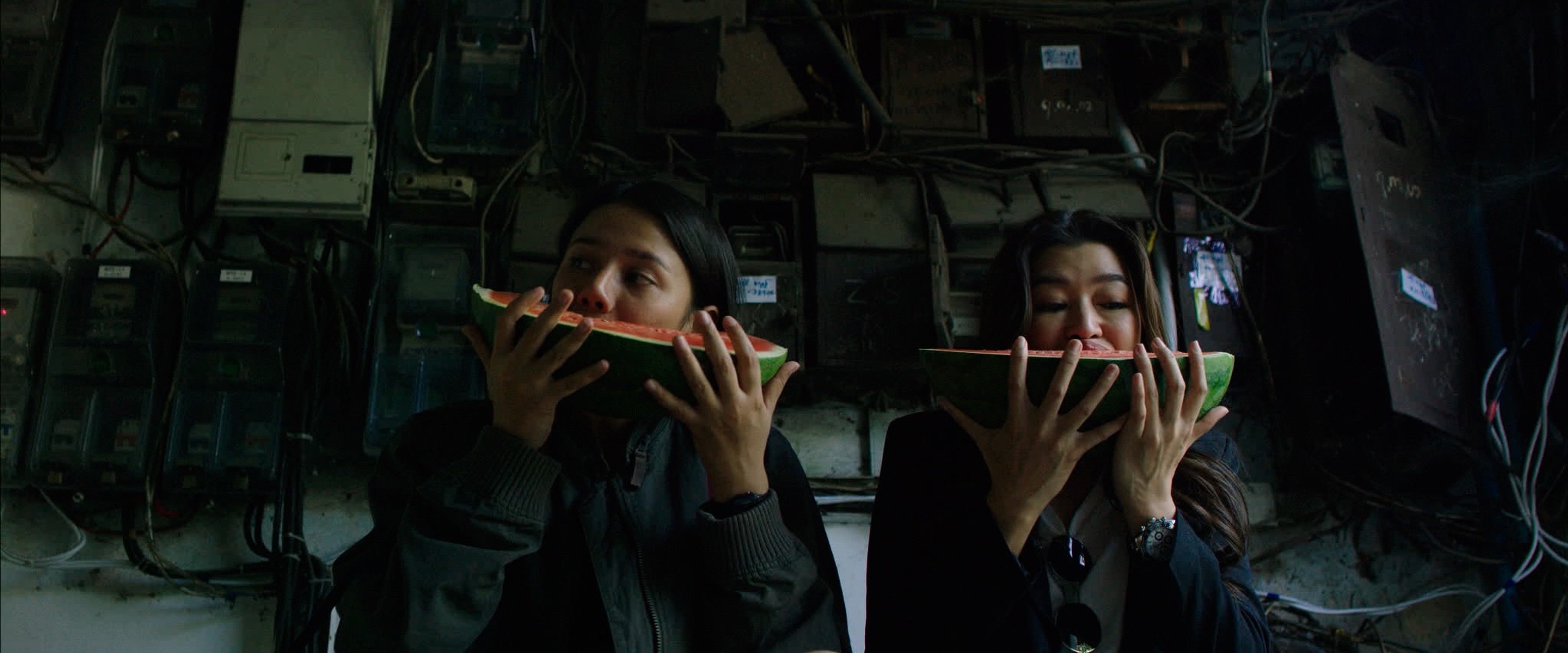 Two women eating slices of watermelon together in a dim industrial-looking space lined with electrical boxes and wiring.