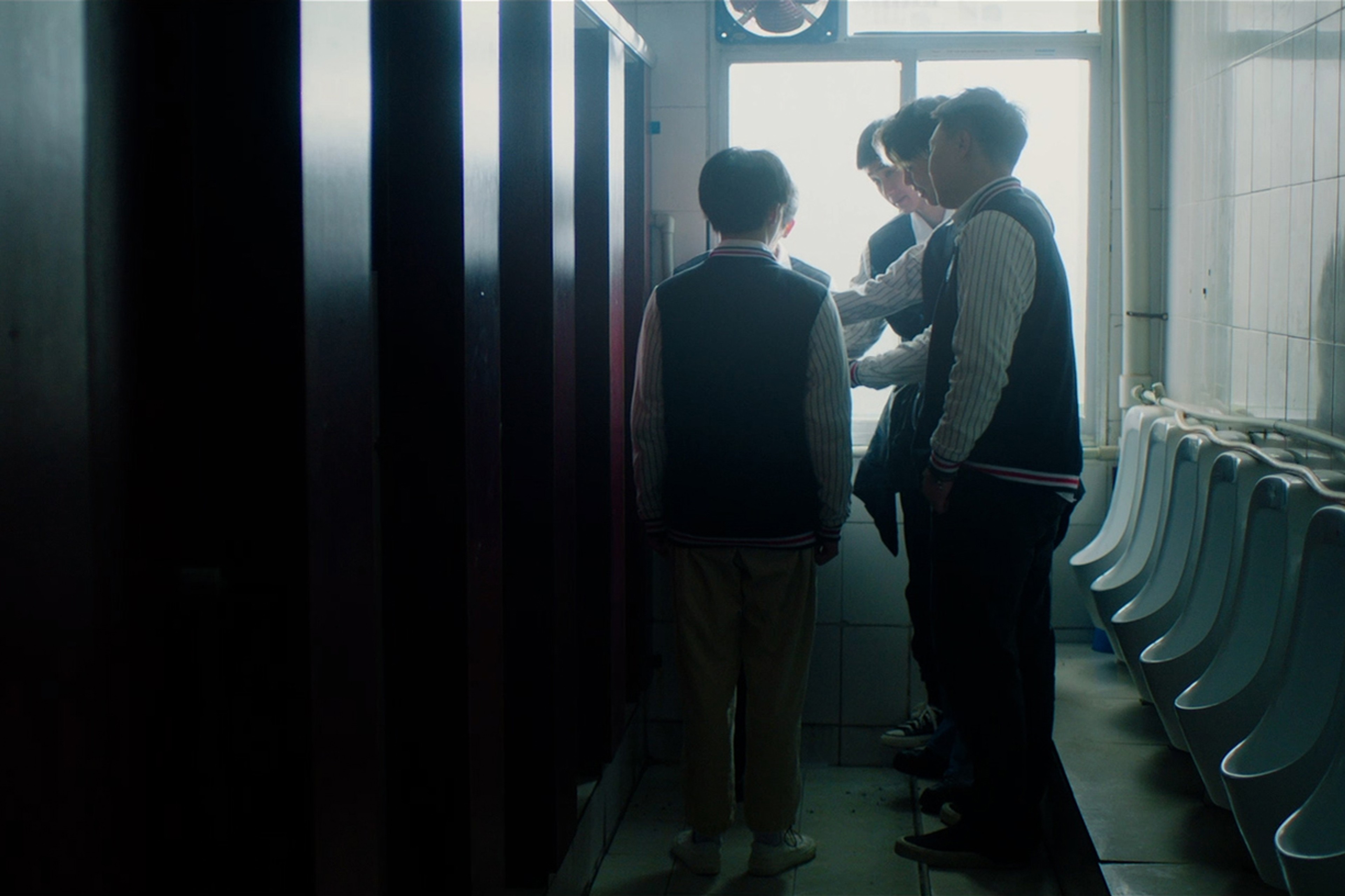 A group of young men gathered in a public restroom near a row of urinals.