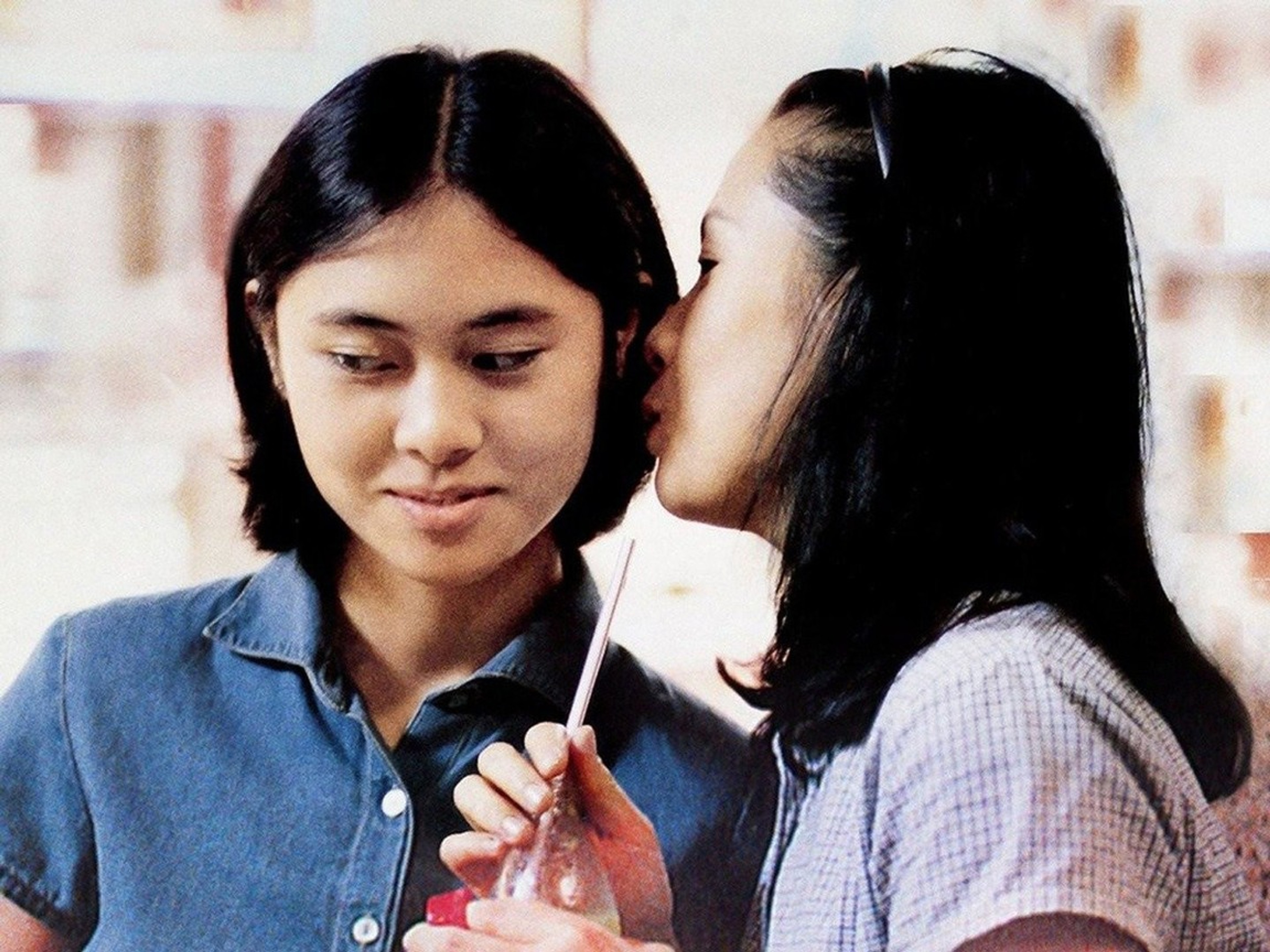 One woman leaning in to whisper into another woman’s ear as they share a drink.