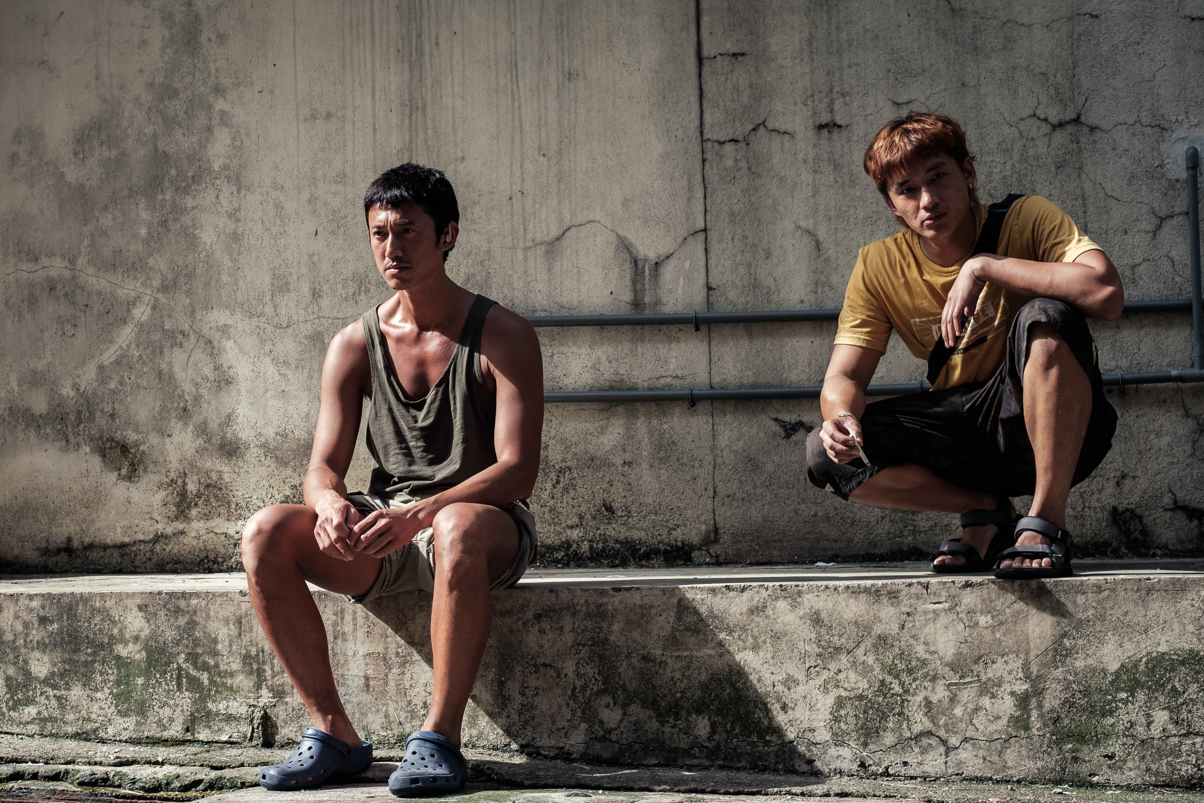 Two young men sitting apart against a weathered concrete wall in bright sunlight.