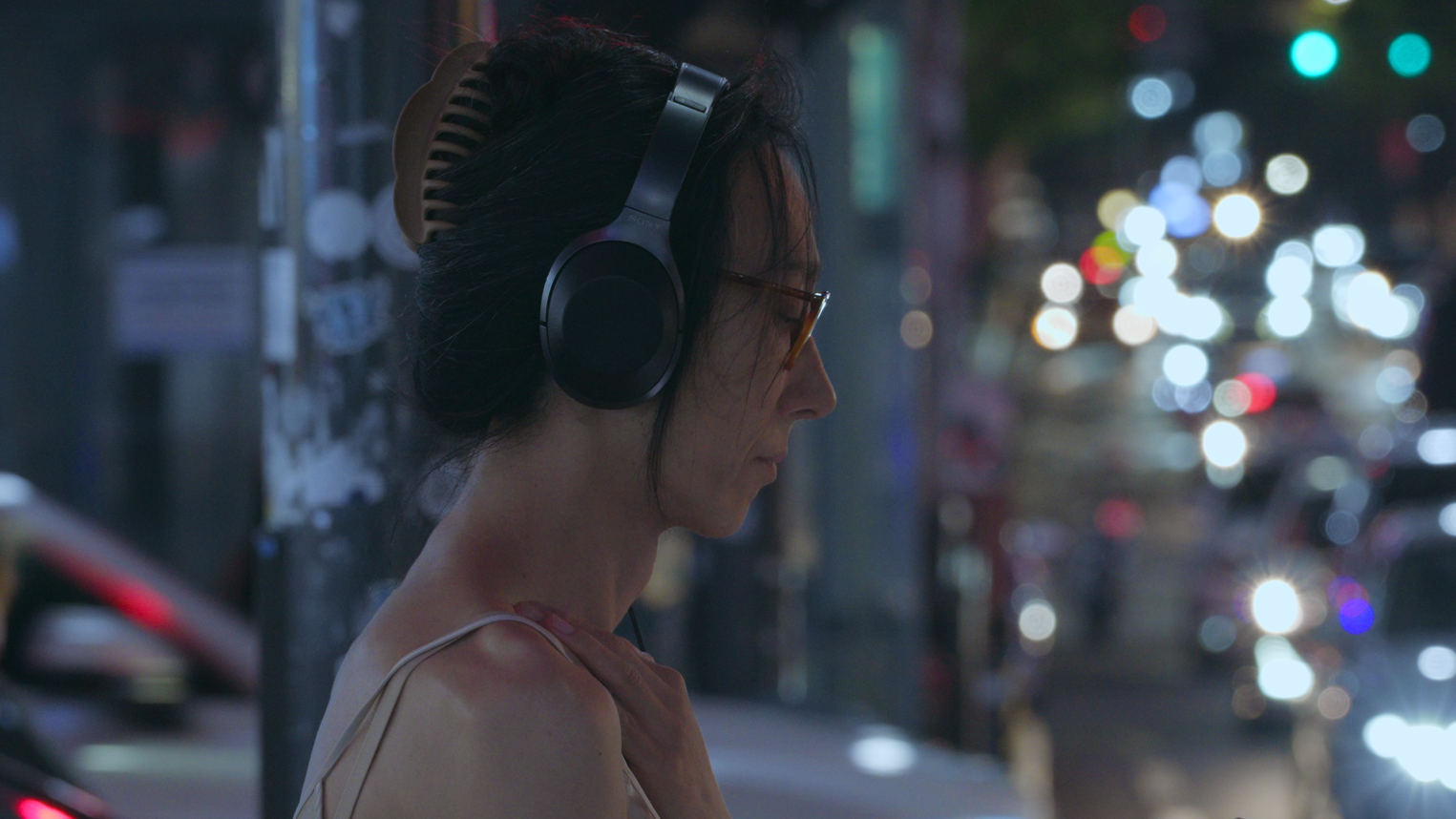 A persom wearing headphones standing on a city street at night, with blurred traffic lights in the background.