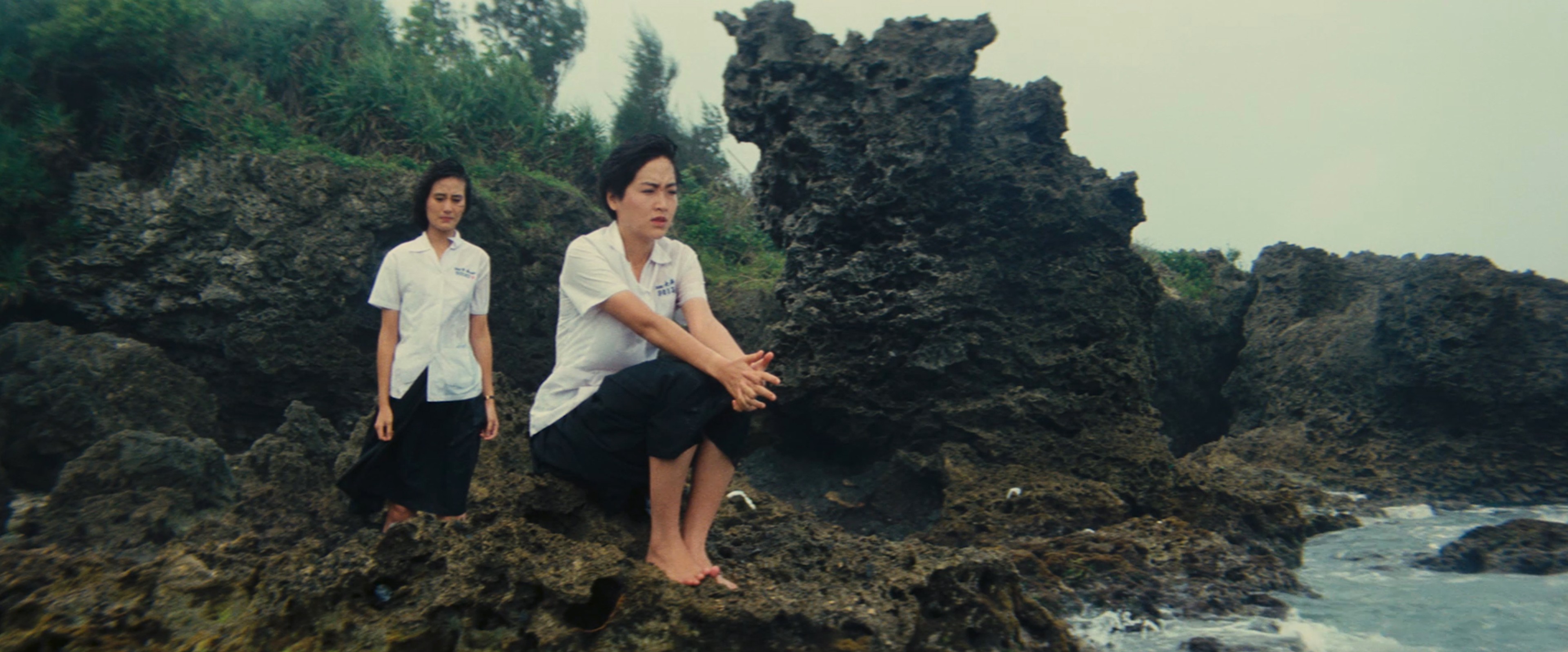 Two women in white shirts standing on a rocky shoreline, one seated barefoot while the other stands behind her.