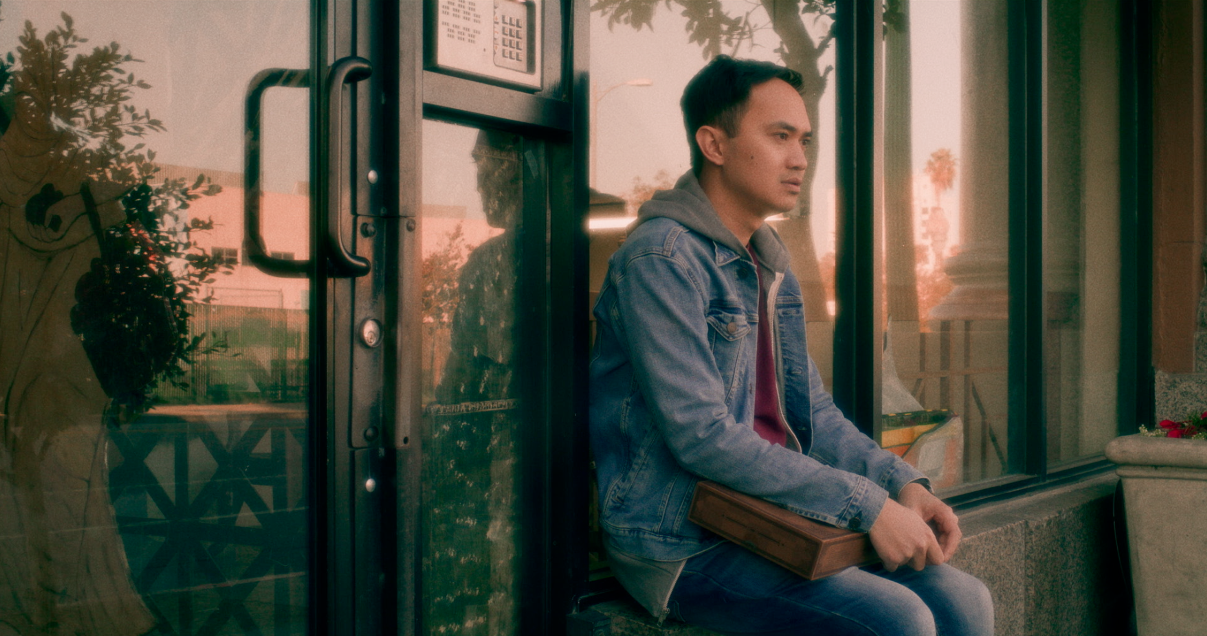 A man sitting outside a storefront, holding a rectangular case on his lap.