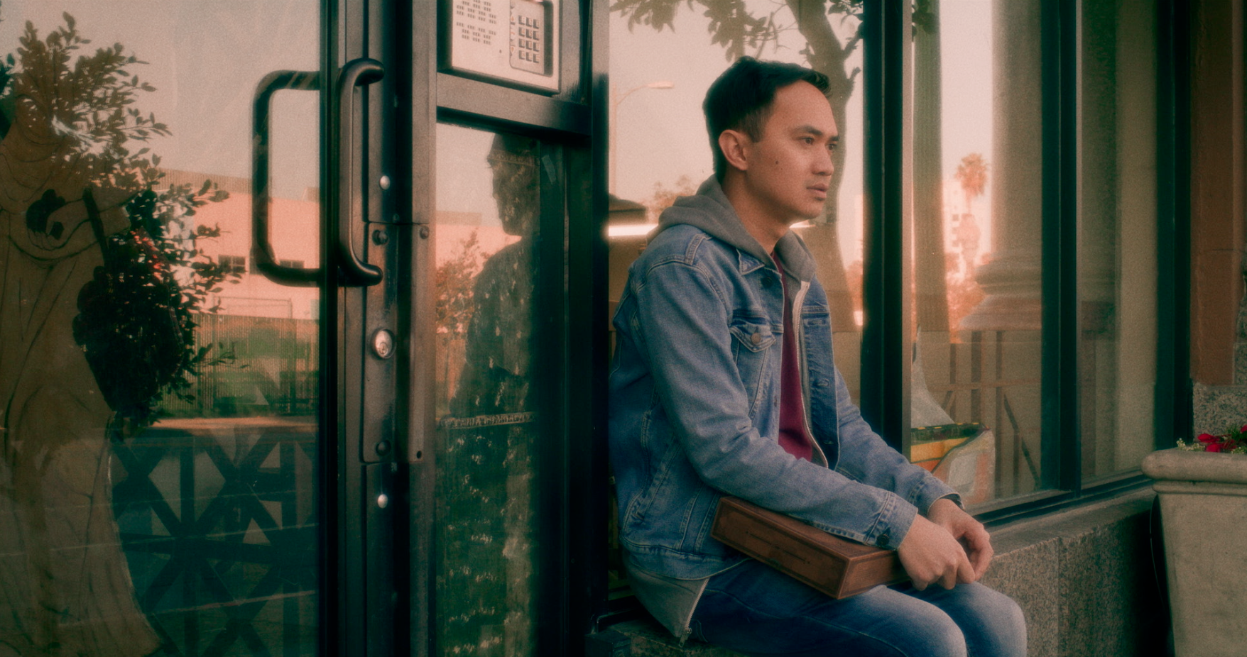 A man sitting outside a storefront, holding a rectangular case on his lap.