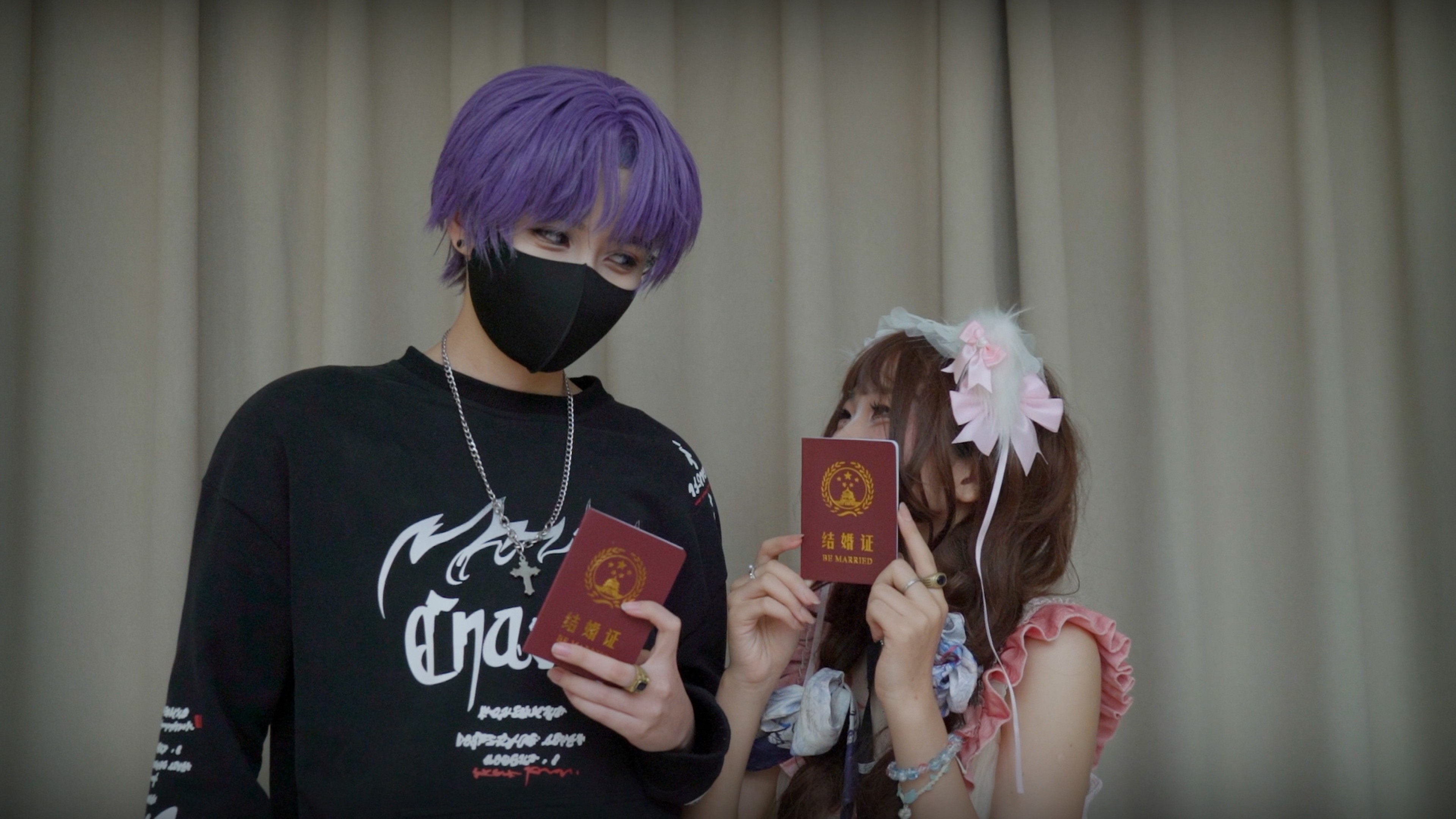Two people in colourful alternative fashion hold up red marriage certificates, standing together in front of a curtain.