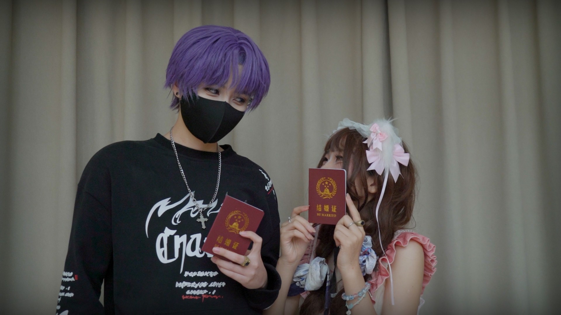 Two people in colourful alternative fashion hold up red marriage certificates, standing together in front of a curtain.