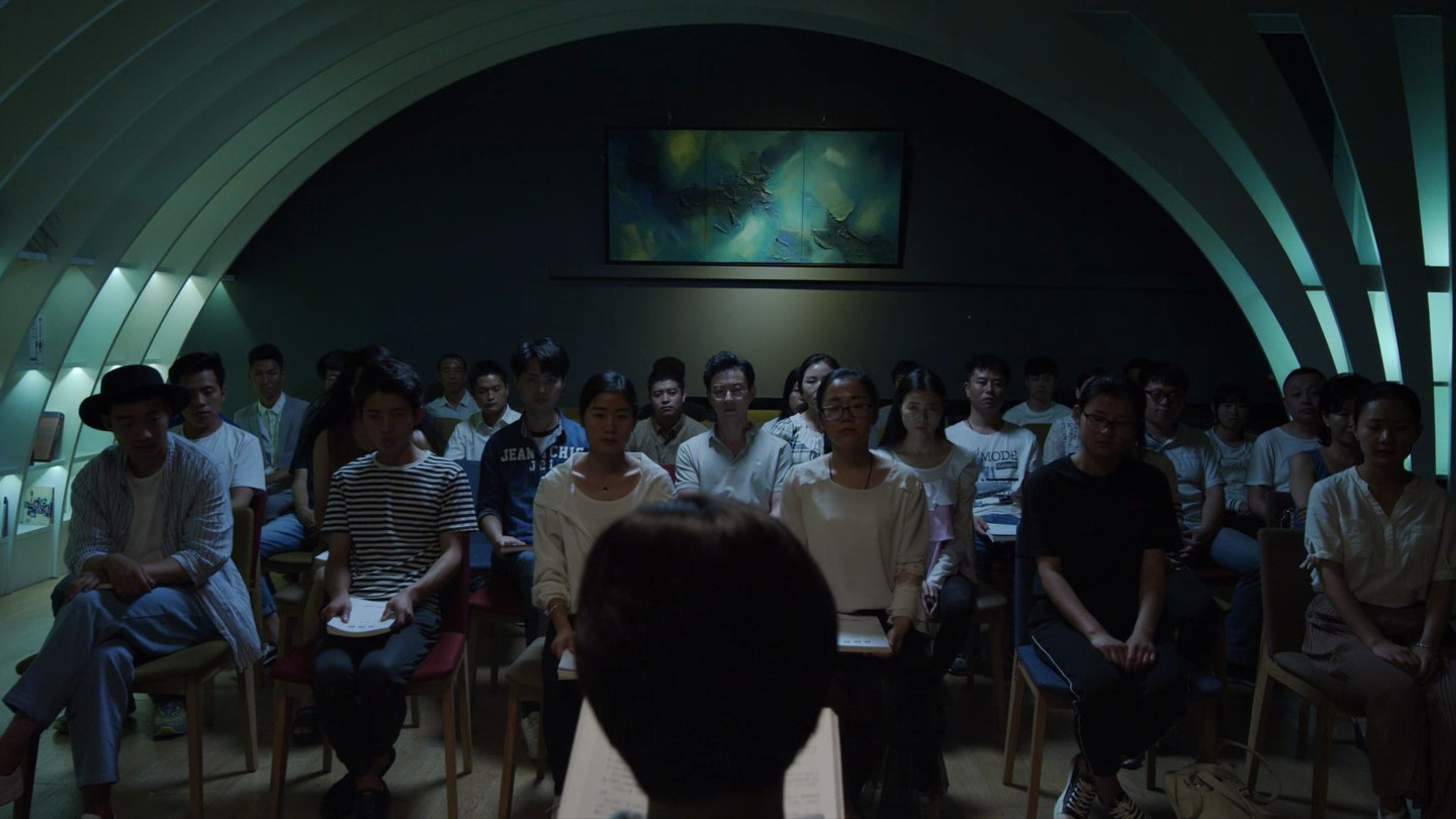 An audience seated in rows inside a dimly lit arched room, viewed from behind a speaker at the front.