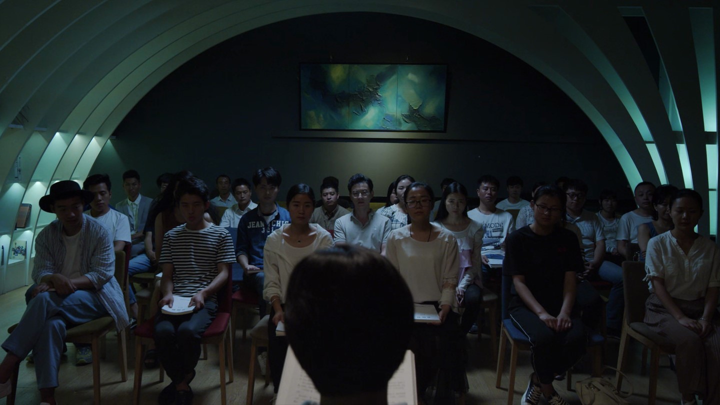 An audience seated in rows inside a dimly lit arched room, viewed from behind a speaker at the front.