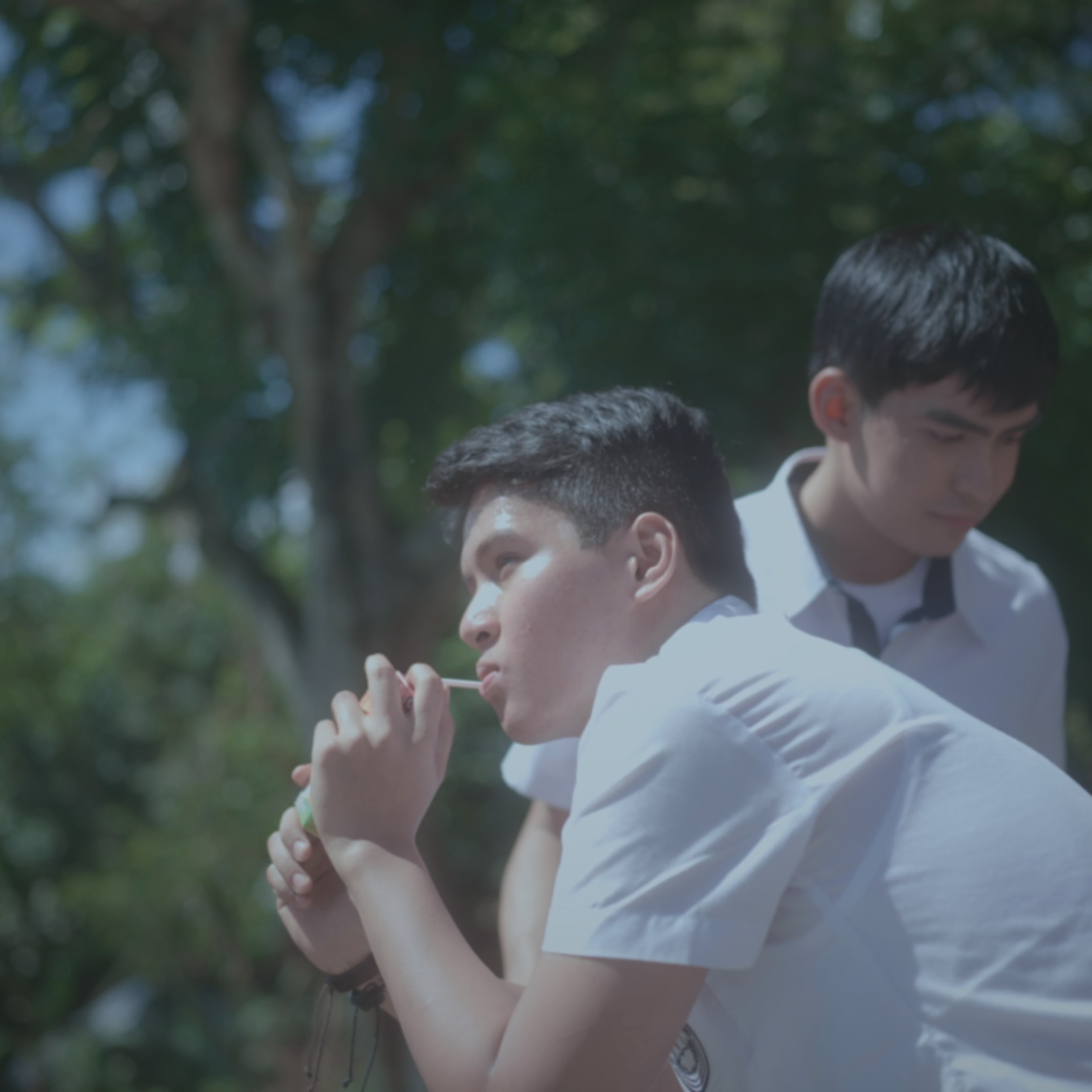 Two young men in school uniforms standing closely together outdoors, one leaning forward with his hands clasped.