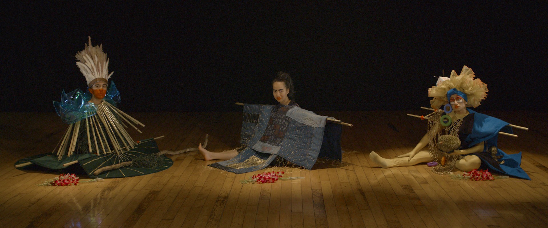 Three figures wearing blue costumes sit on a wooden floor.