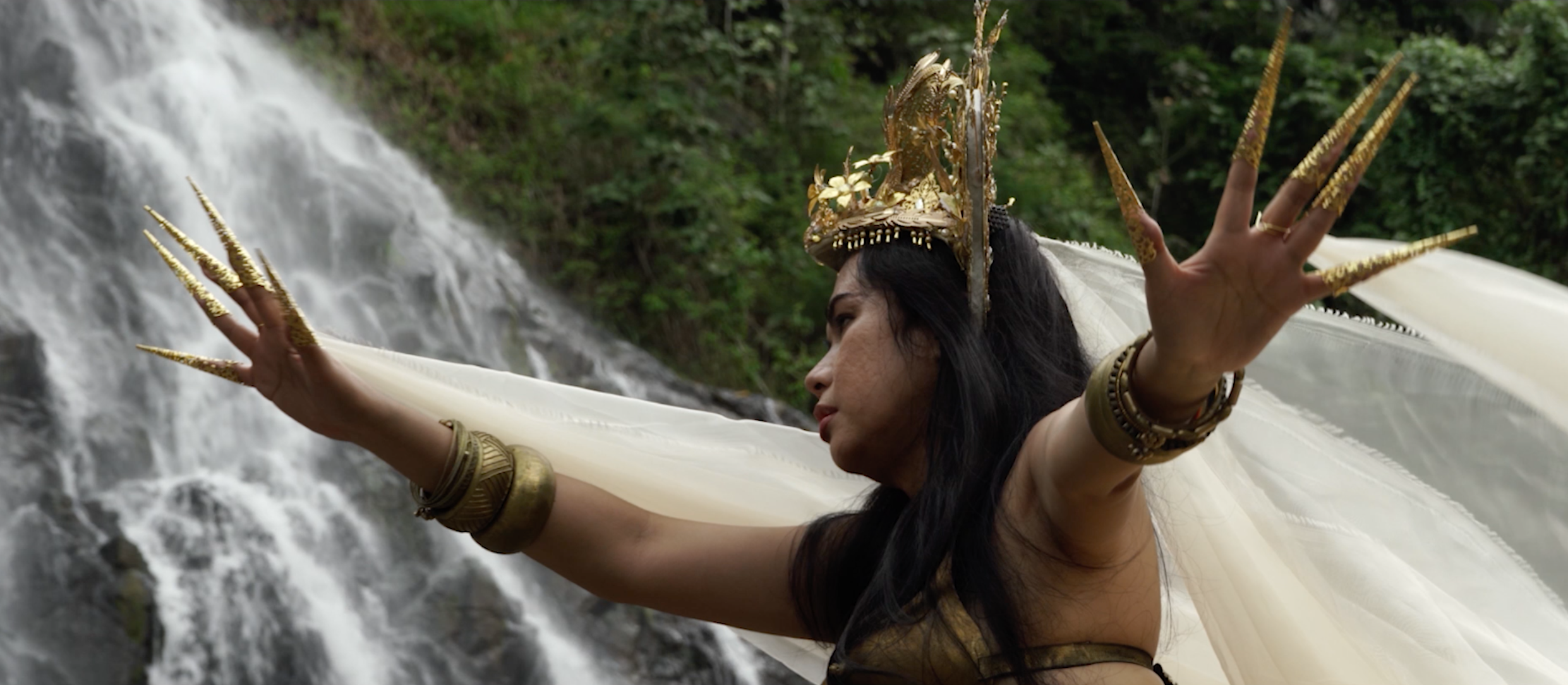 A woman wearing flowing white robes and spiky golden nails stands in front of a waterfall.