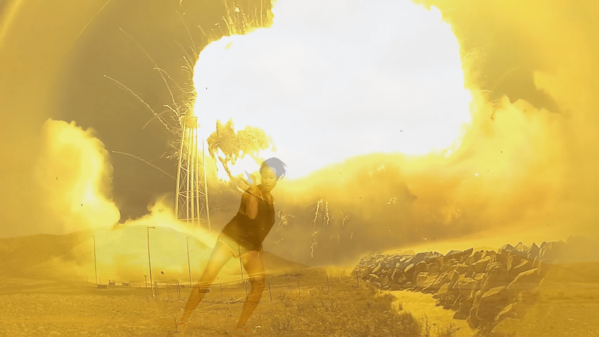 A person standing in a barren landscape as a massive explosion erupts behind them.