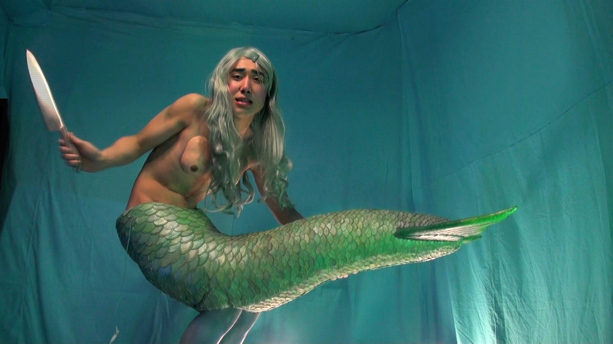 A mermaid-like figure with long silver hair holding a large knife against a turquoise backdrop.
