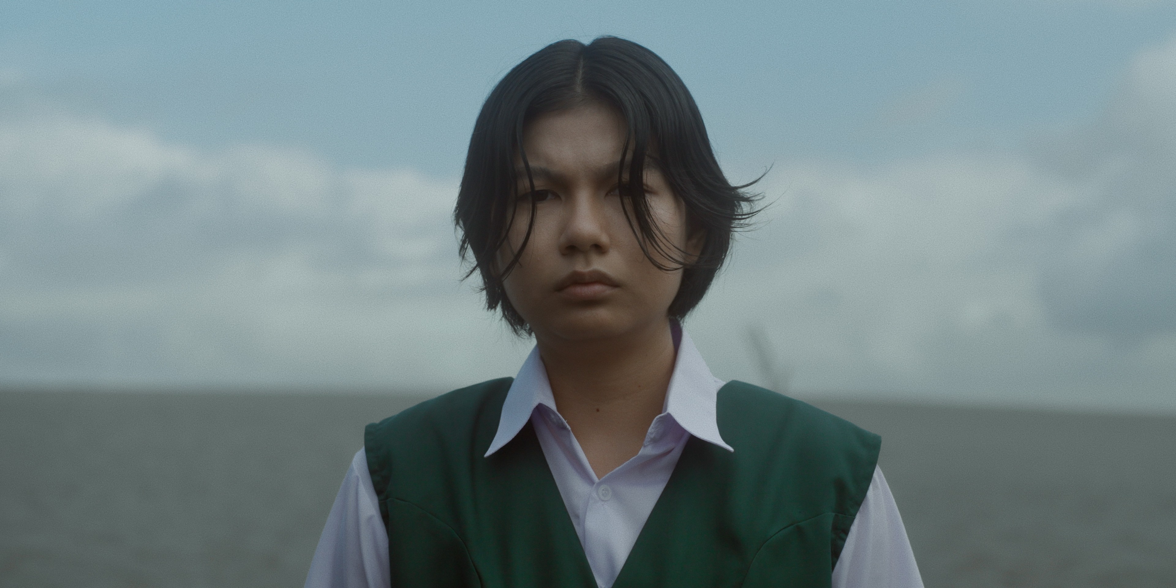 A young person in a green school uniform standing by the sea beneath an overcast sky.