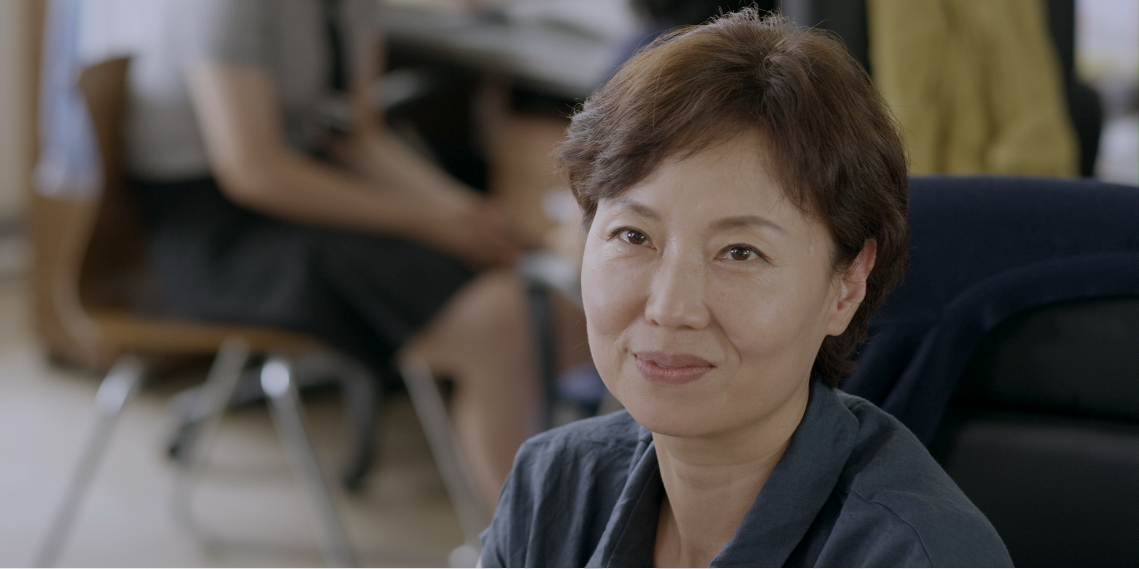 A woman with short hair smiling gently while seated in a bright office or classroom.