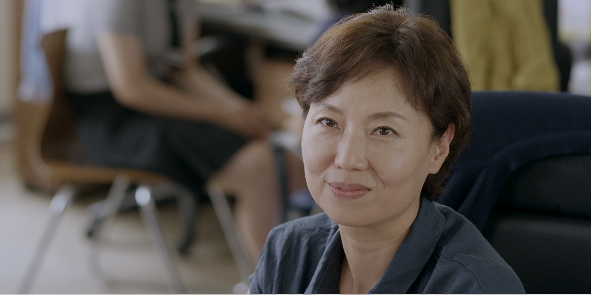 A woman with short hair smiling gently while seated in a bright office or classroom.