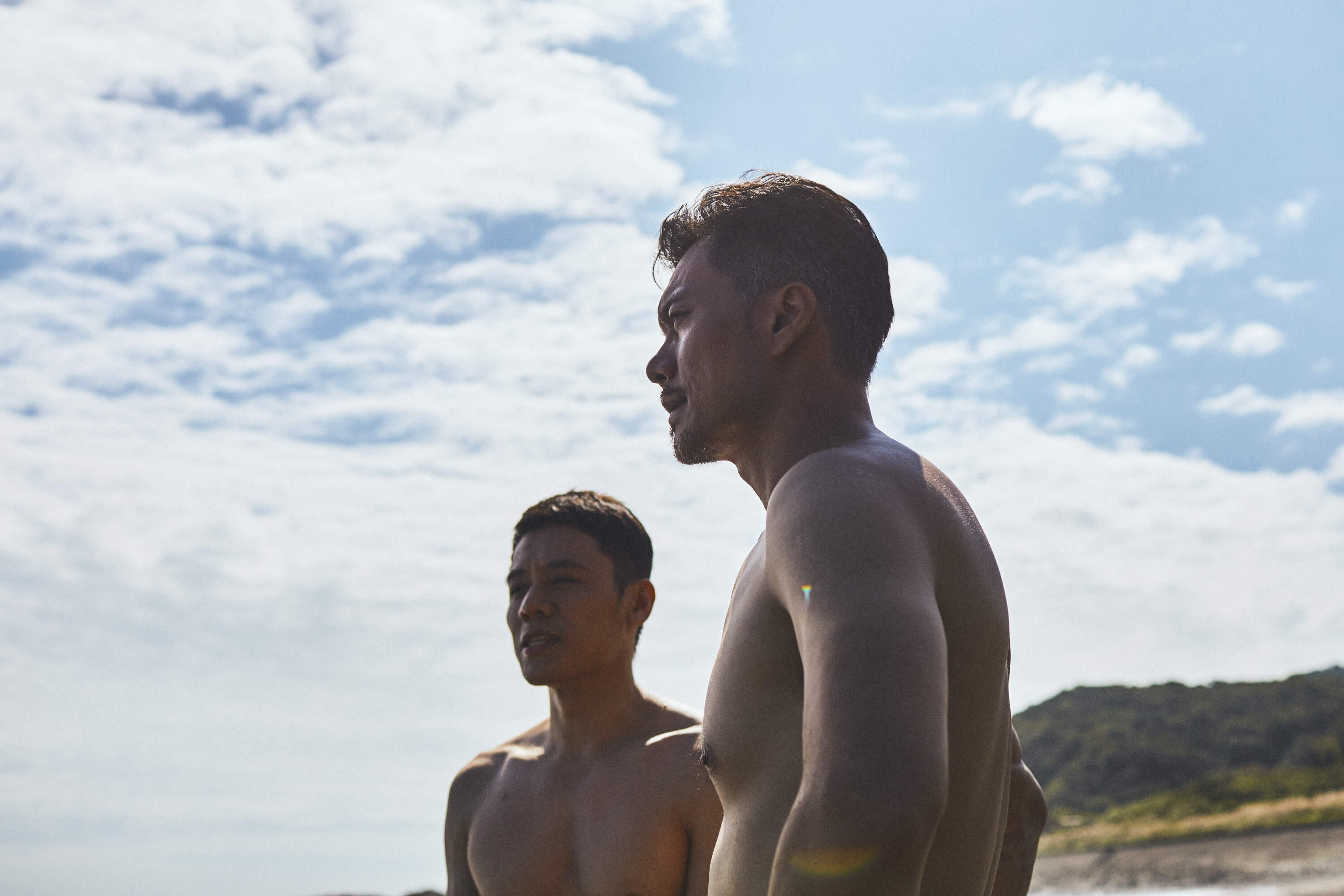 Two shirtless men against a backdrop of a blue sky with clouds.